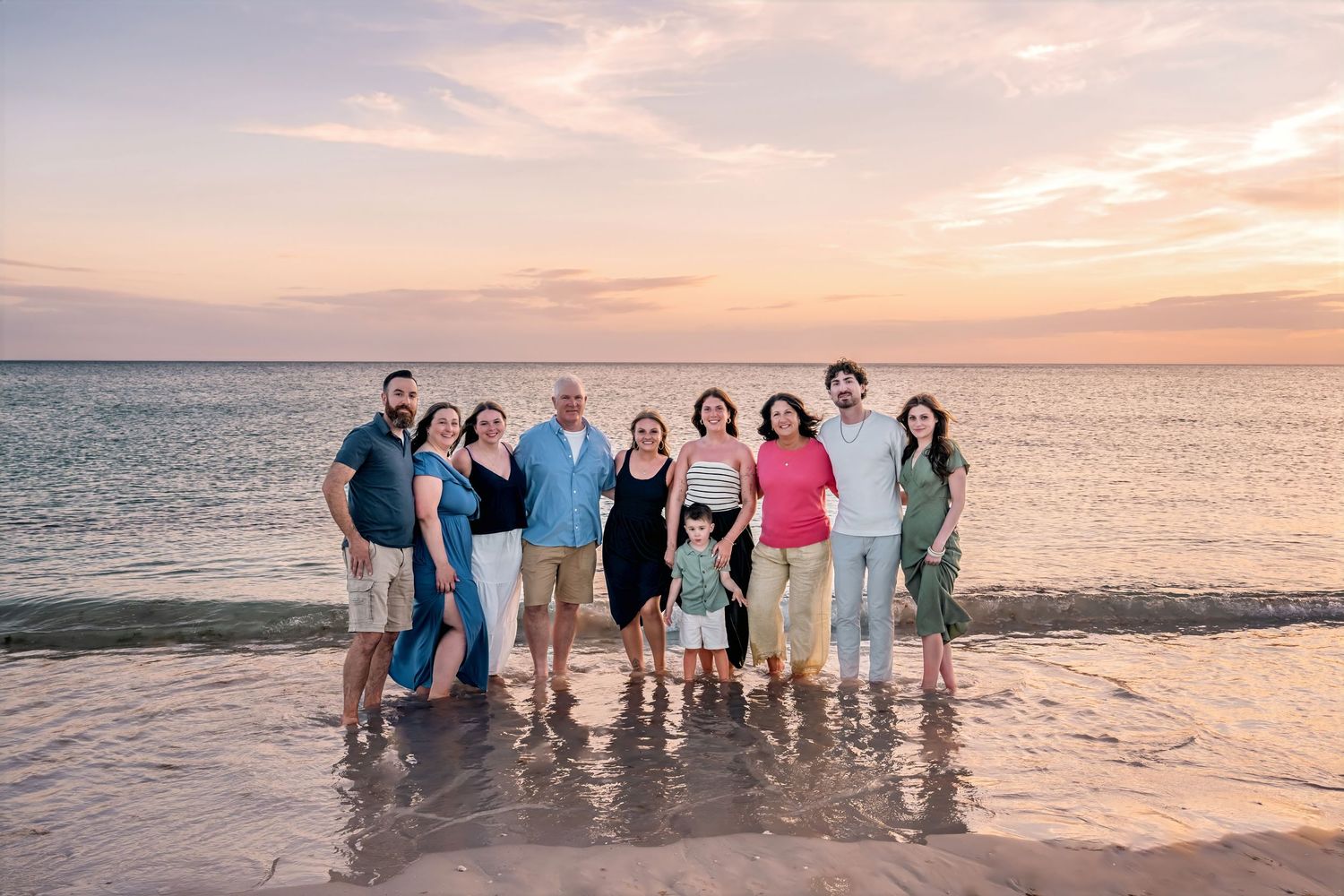 Family group photo on a sandy beach at sunset with pink and orange sky over calm ocean waters.