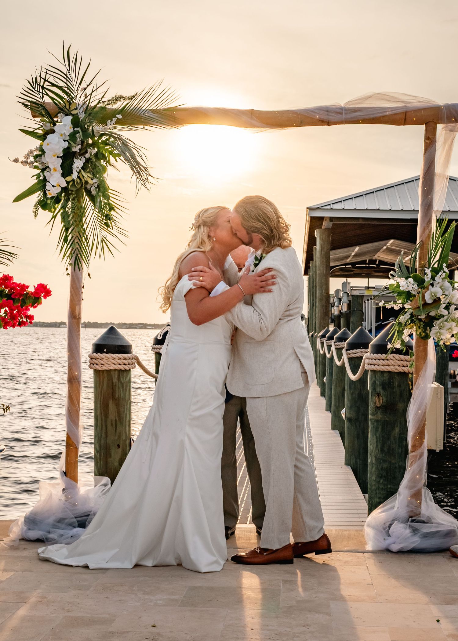 Bride and groom share first kiss at sunset waterfront ceremony under a floral arch on a dock.