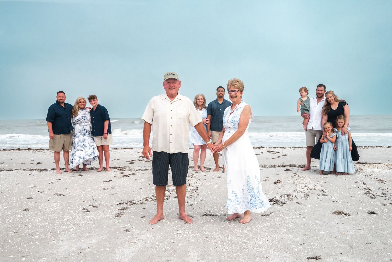 Couple stands in foreground on beach with extended family posed behind them near ocean waves.
