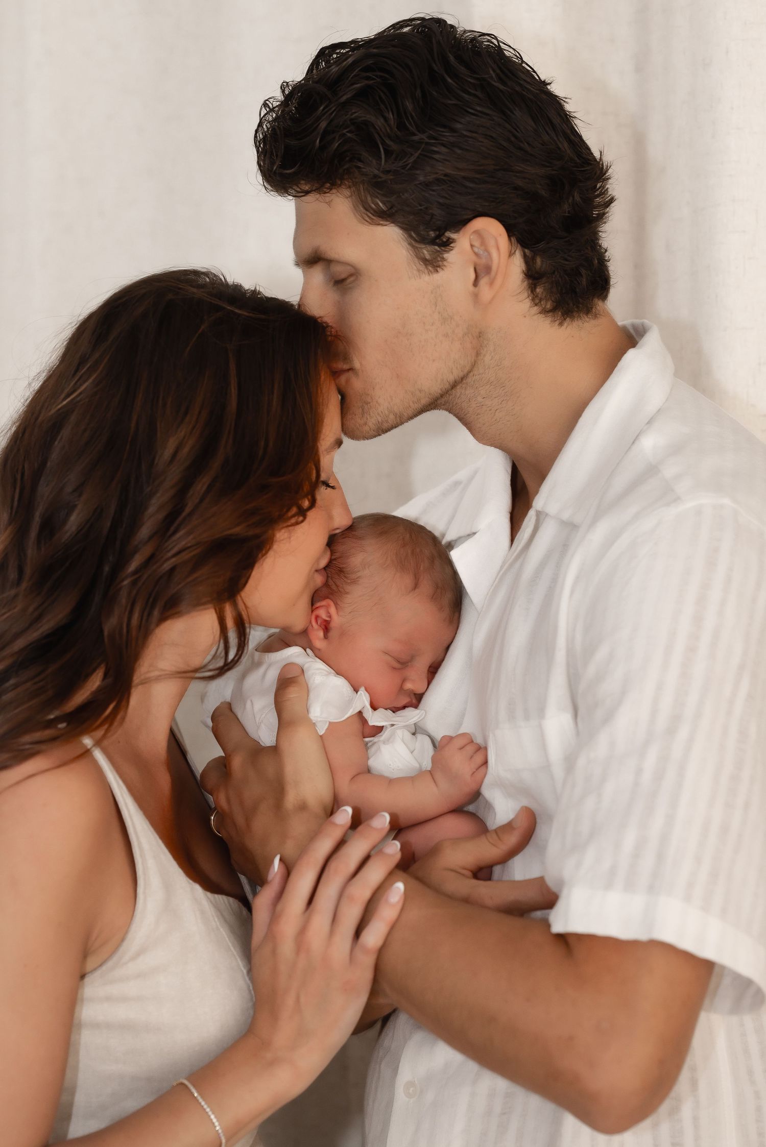 A couple tenderly kisses while cradling their sleeping newborn baby dressed in white.