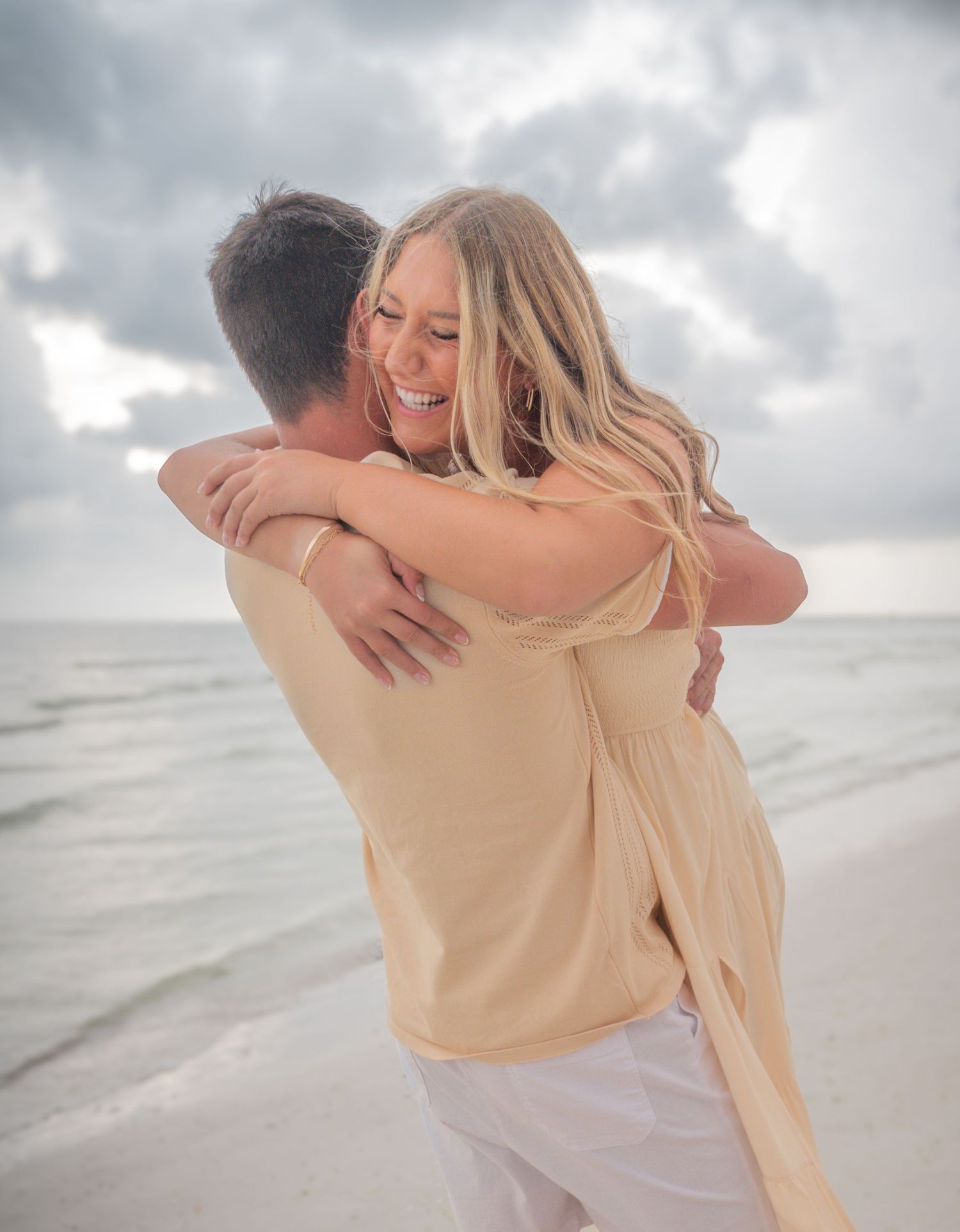 Joyful woman in a yellow dress hugs her partner on a cloudy beach shoreline.