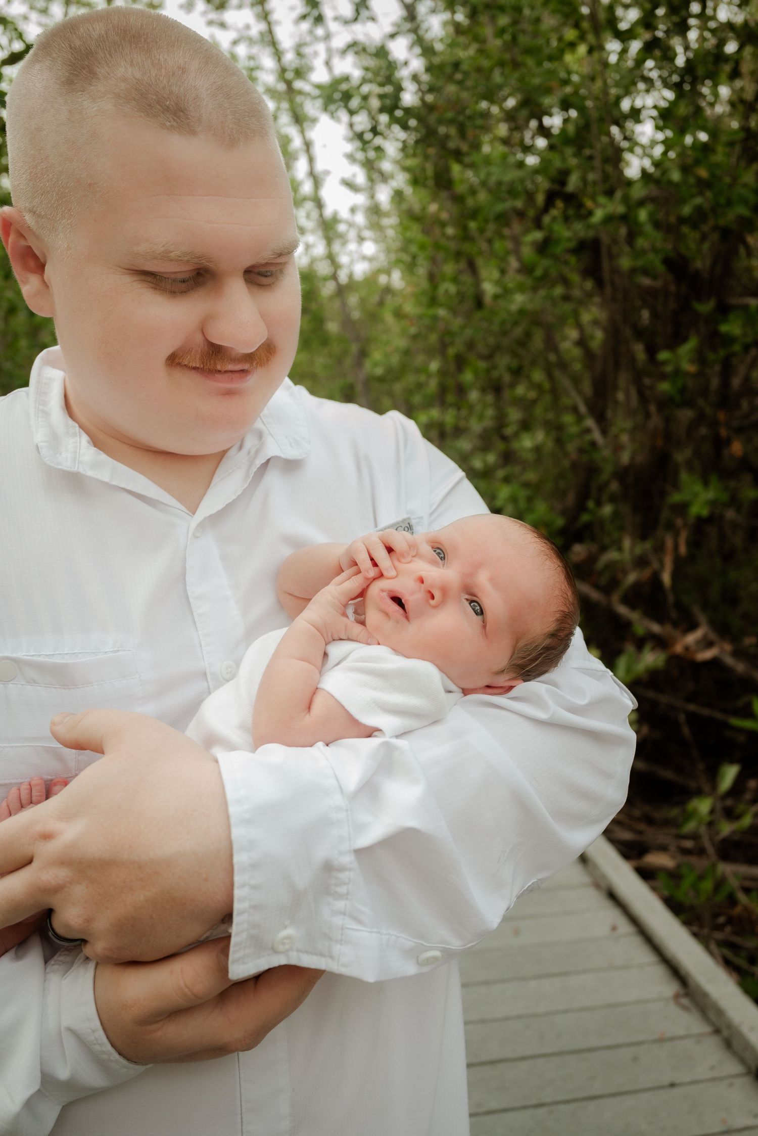New father in white shirt tenderly holding smiling newborn baby outdoors on wooden path.