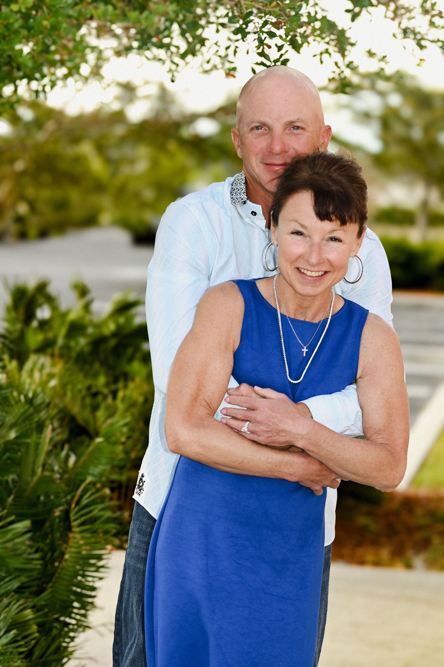 Smiling couple poses outdoors, bald man hugs woman in blue dress from behind.
