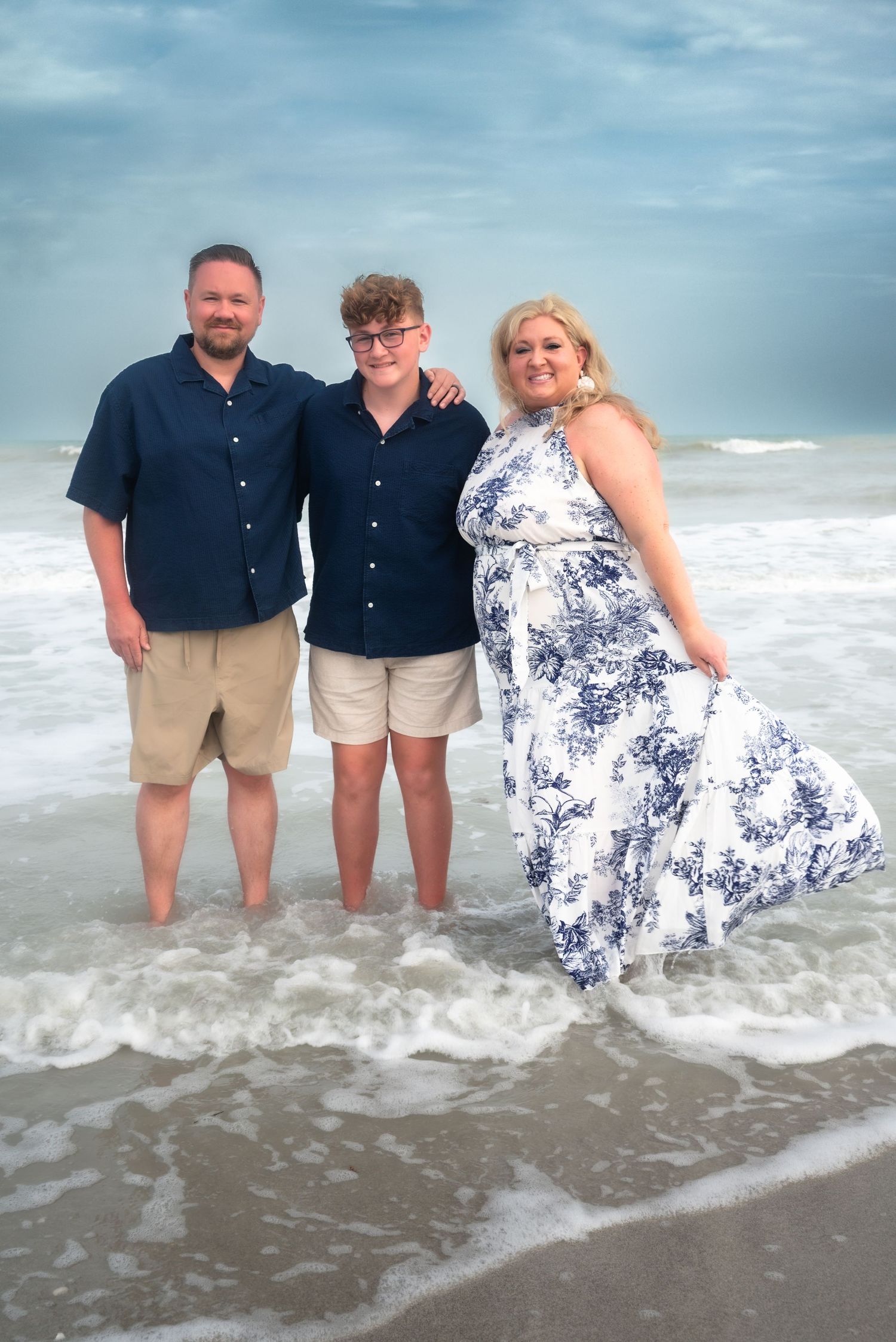 A family of three stands smiling in ocean waves at the beach.