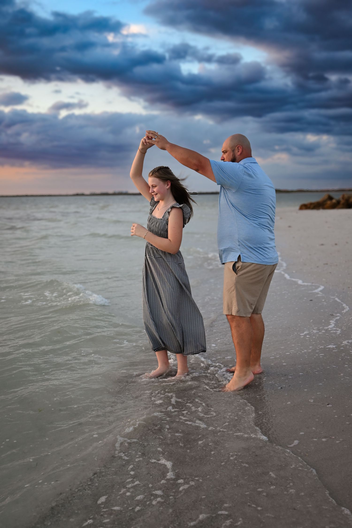 Romantic couple dancing in ocean waves at sunset, man spinning woman in a gray dress under dramatic cloudy skies.