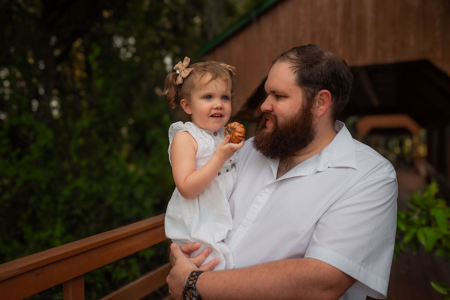 Bearded father in white shirt holds smiling toddler daughter outdoors near wooden covered bridge.
