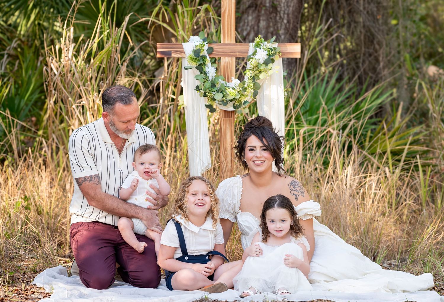 Family of five sitting on white blanket in front of a wooden cross surrounded by tall grass outdoors.