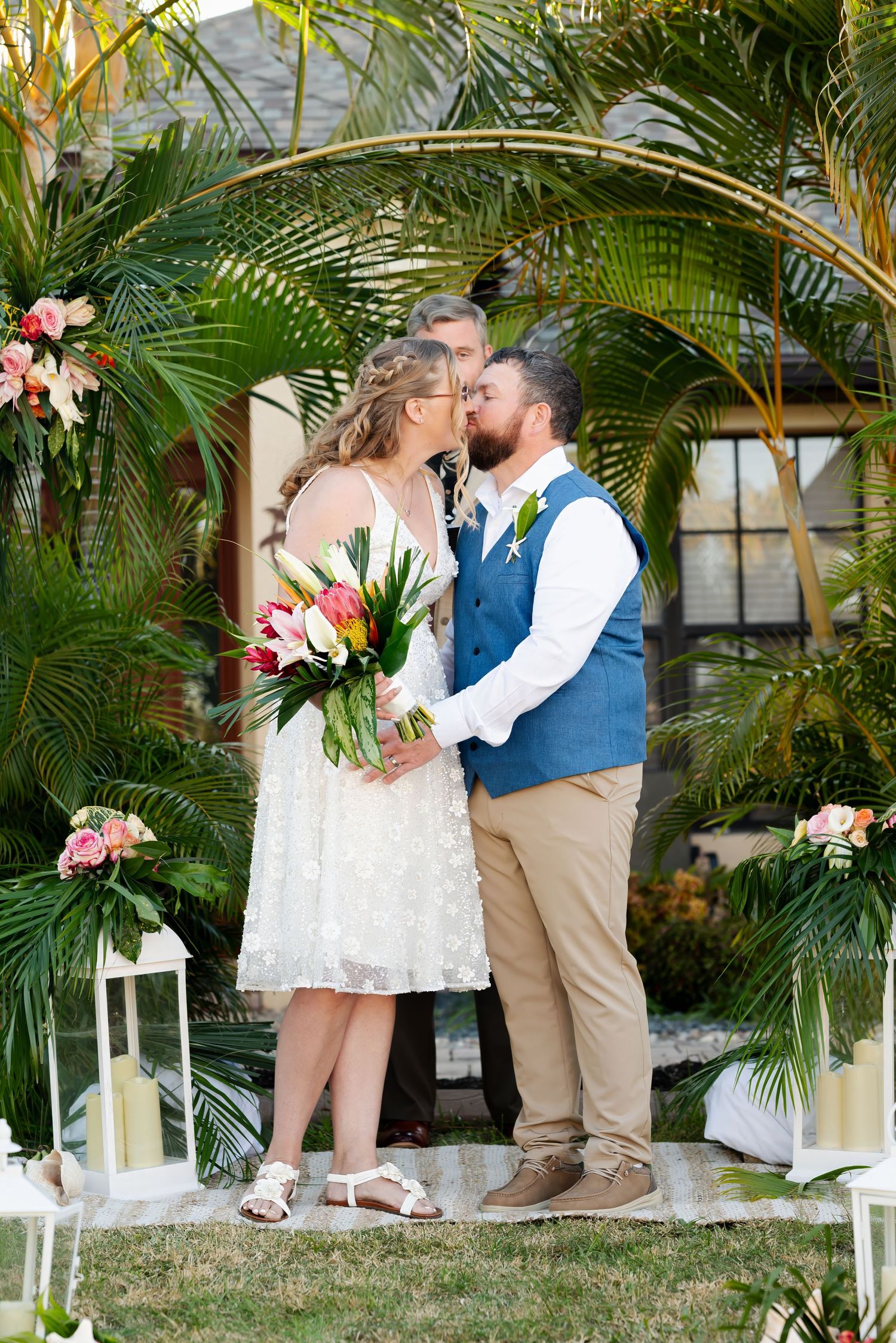 Bride and groom sharing first kiss under a golden arch adorned with tropical flowers.