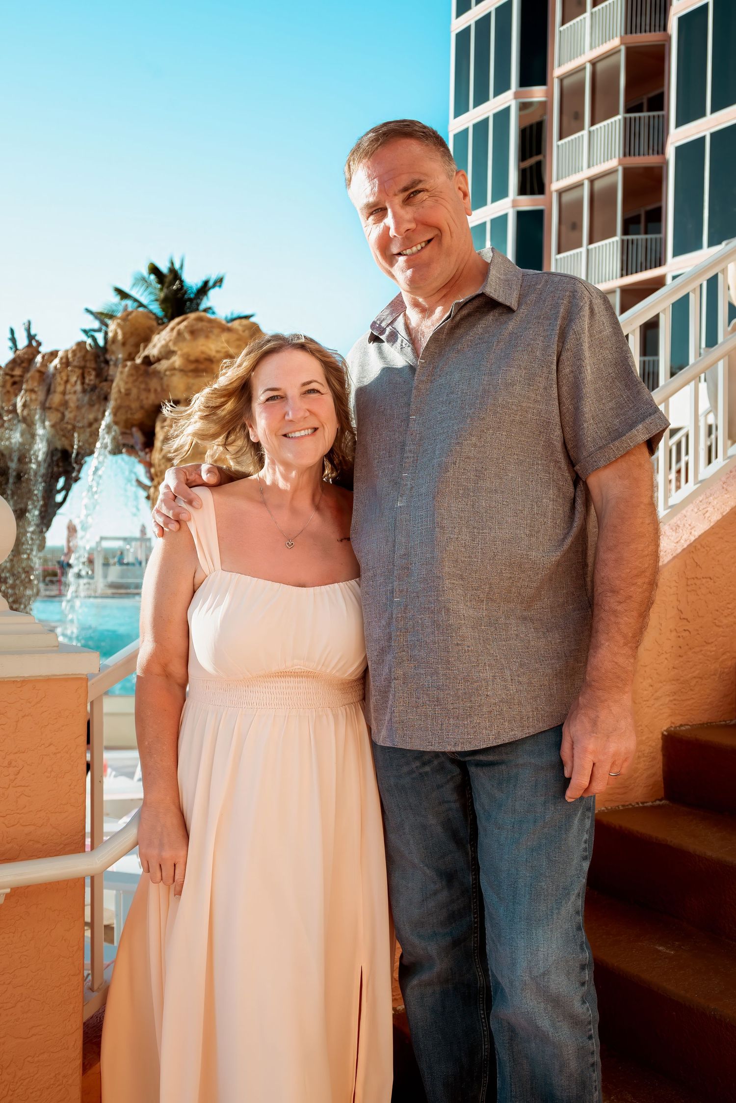 Smiling couple posing outdoors near a fountain and resort building on a sunny day.