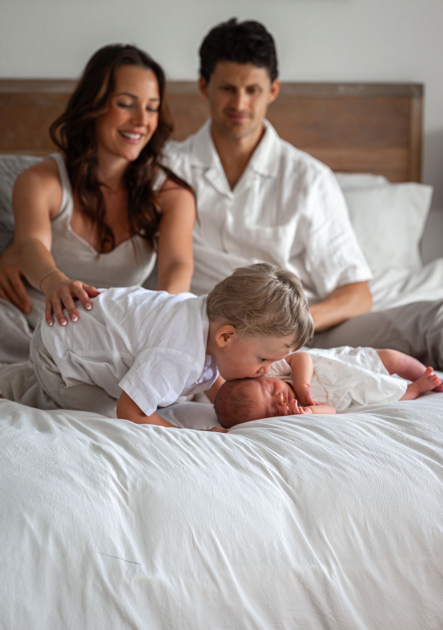 Young boy gently kisses newborn sibling on bed while parents watch lovingly behind them.