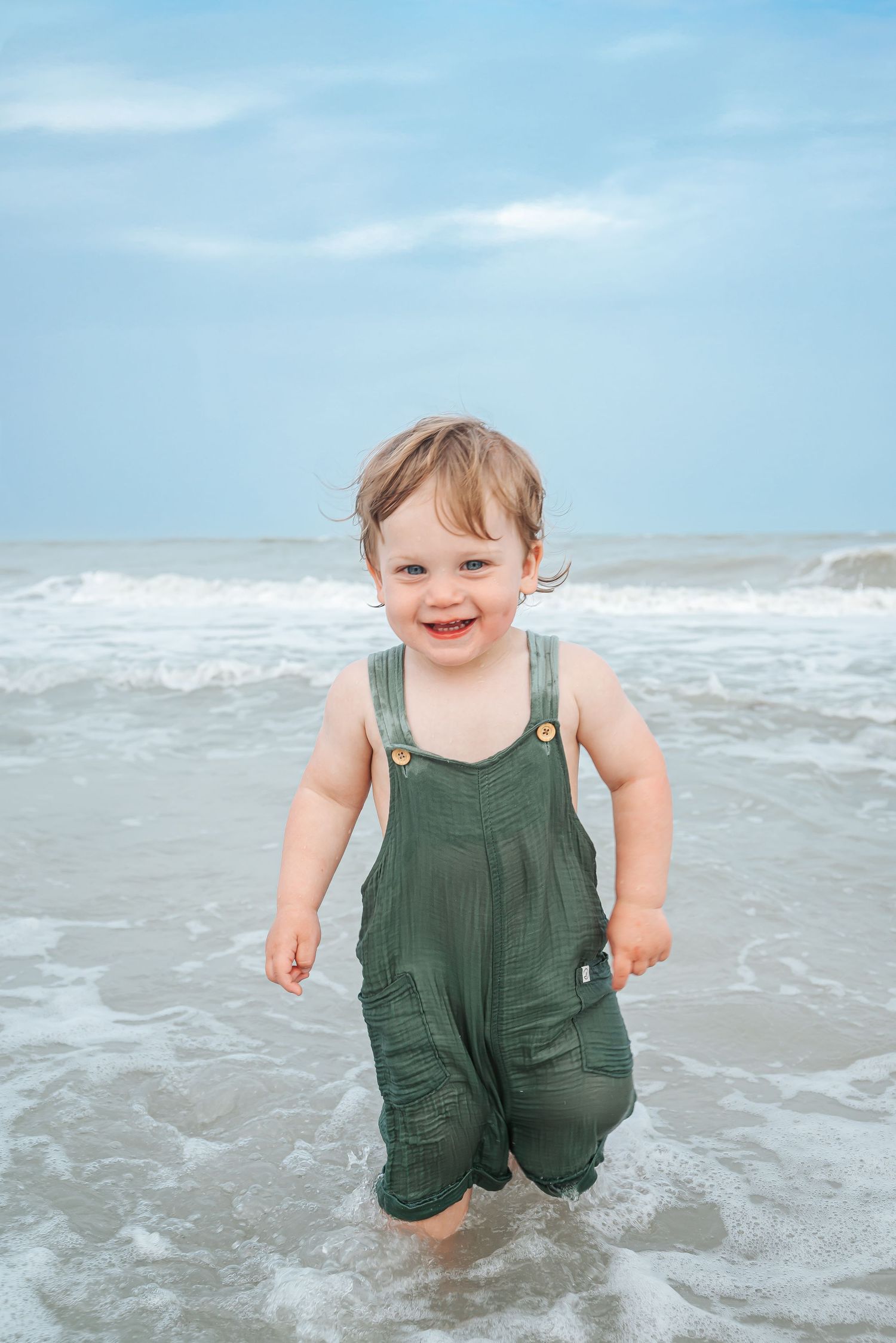 A happy toddler in green overalls splashes in ocean waves on a beach.