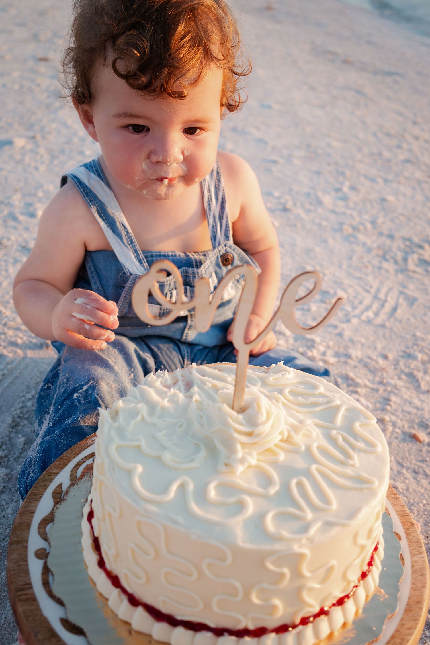 Cute baby in denim overalls celebrating first birthday with white cake on a sandy beach at sunset.
