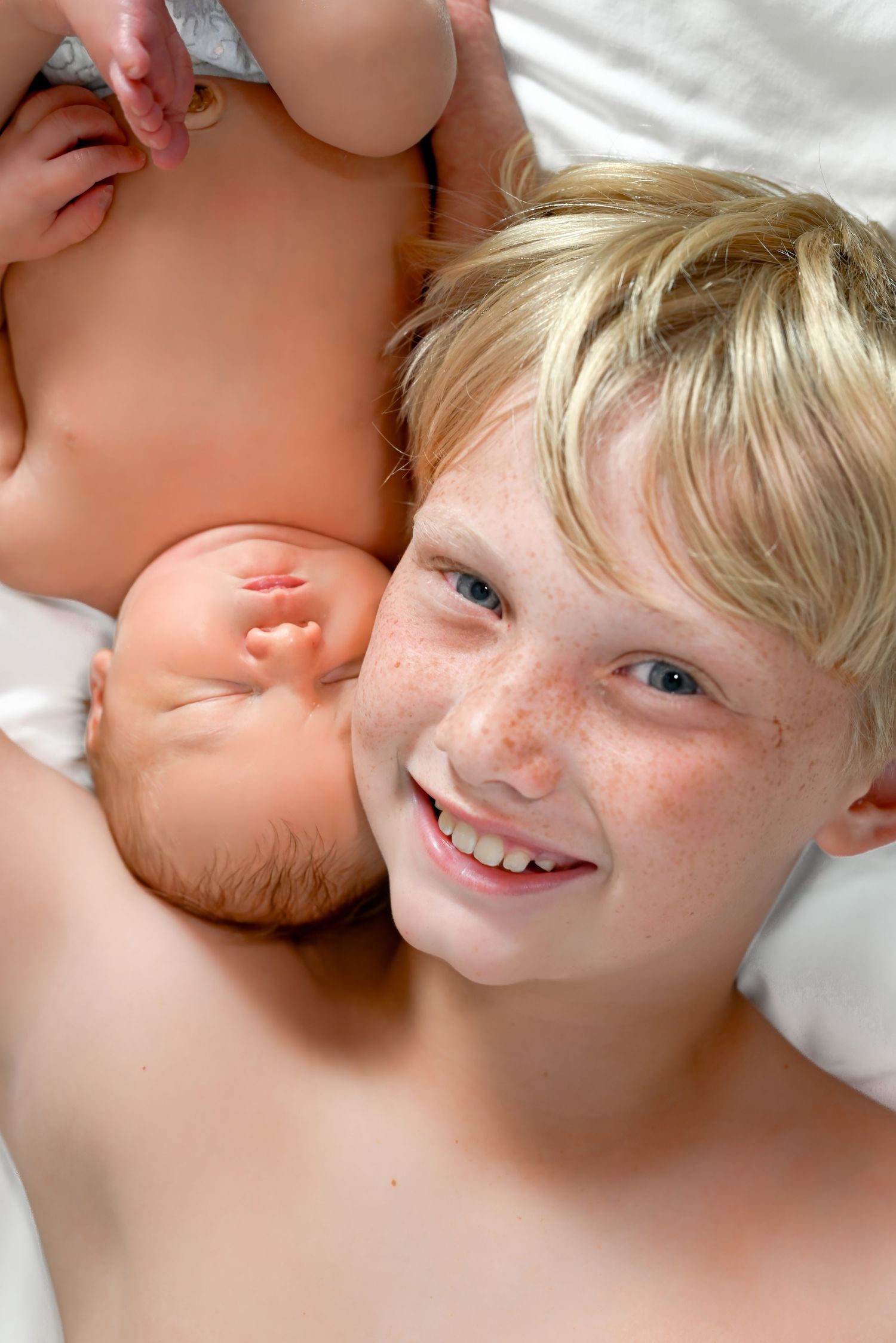 Smiling freckled blonde boy lying next to a sleeping newborn baby on a white bed.