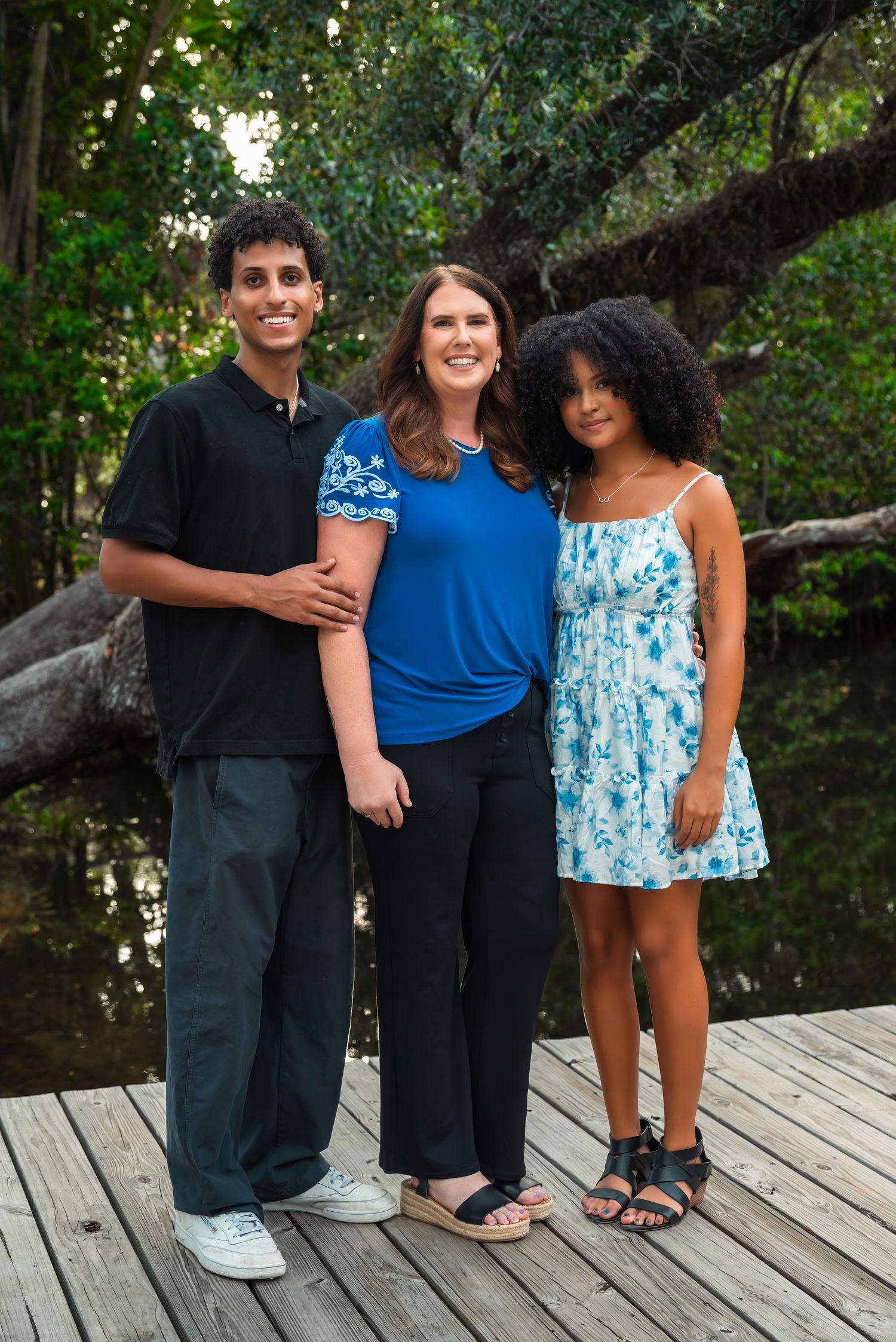 Three people smiling together on a wooden dock surrounded by lush green trees near a tranquil pond.