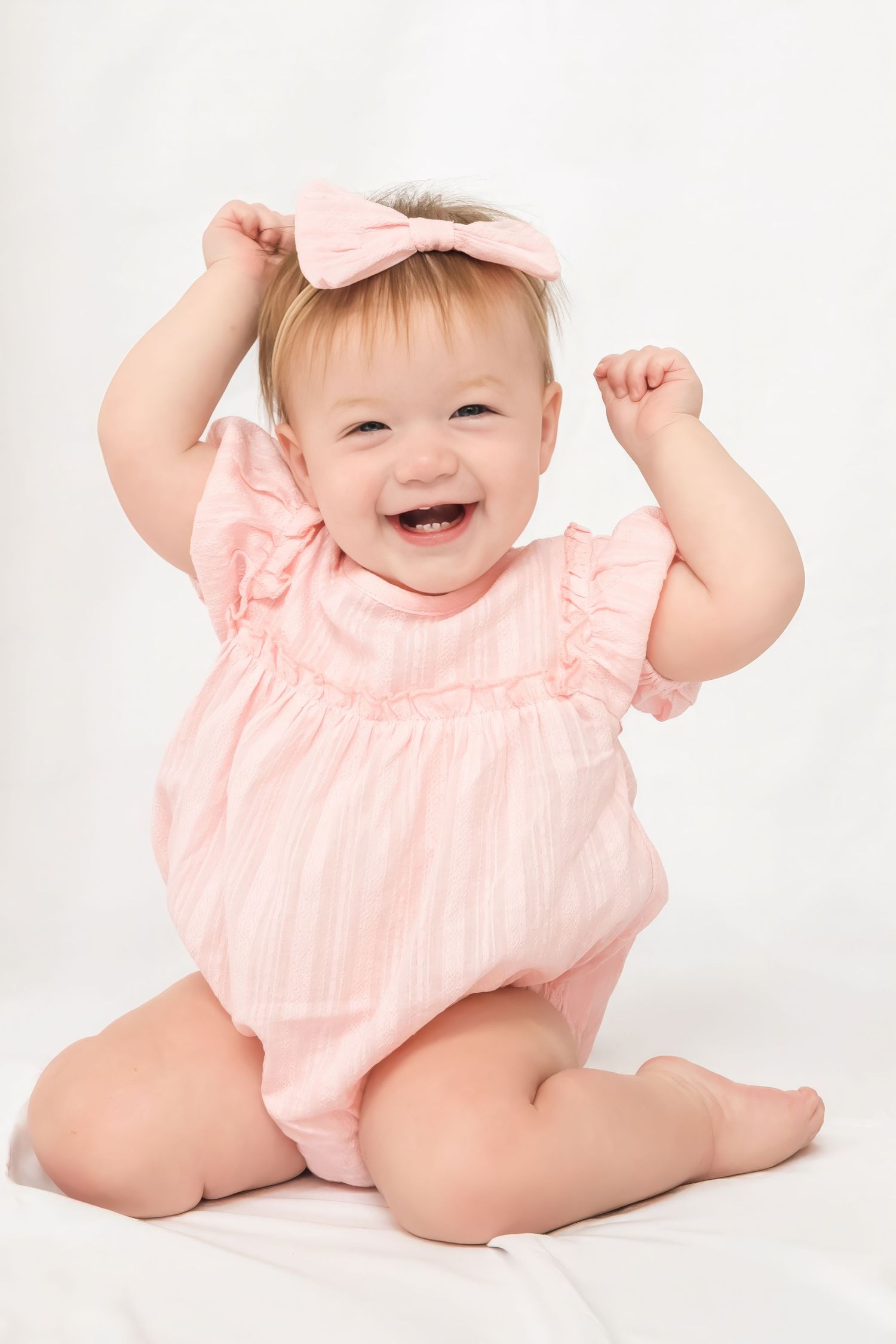 Smiling baby girl in pink romper and bow headband sitting up with arms raised on white background.