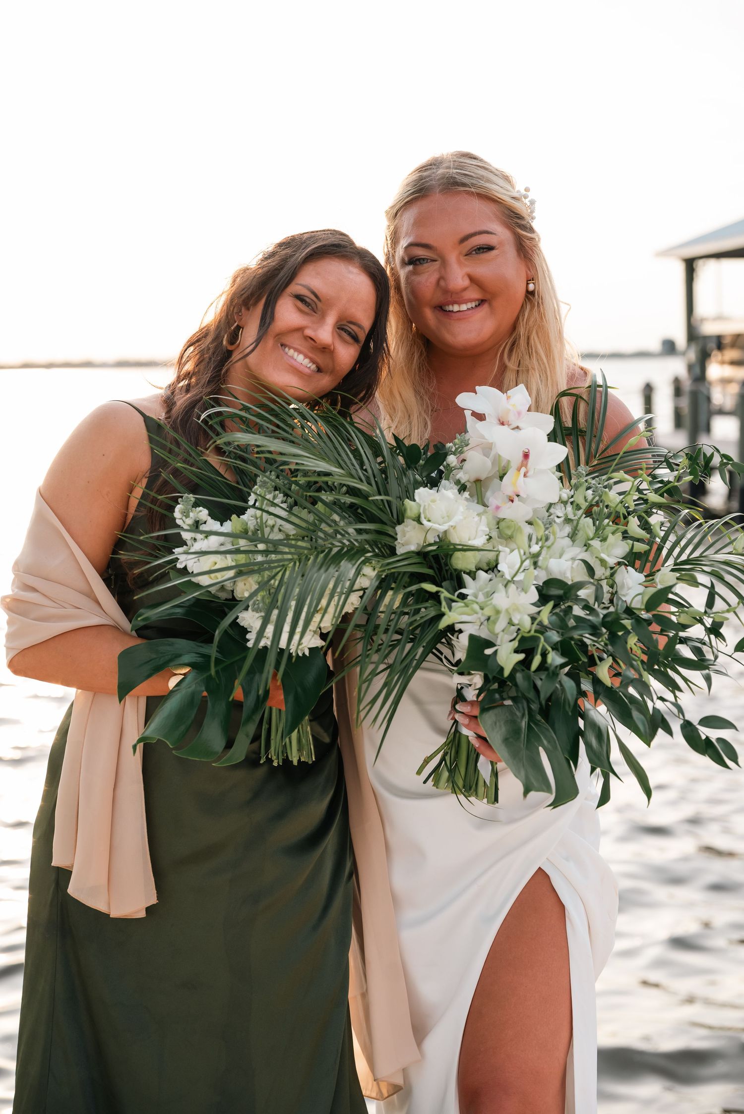 Bride in white dress holding tropical bouquet poses with bridesmaid in olive green dress by waterfront.