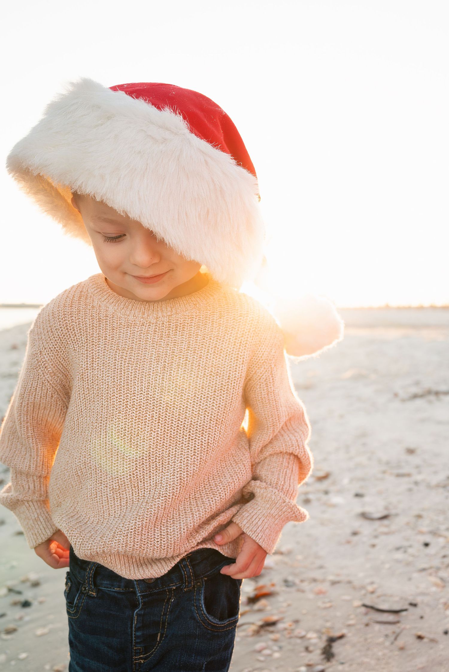 Toddler in beige sweater and Santa hat standing on a sunny beach with lens flare.
