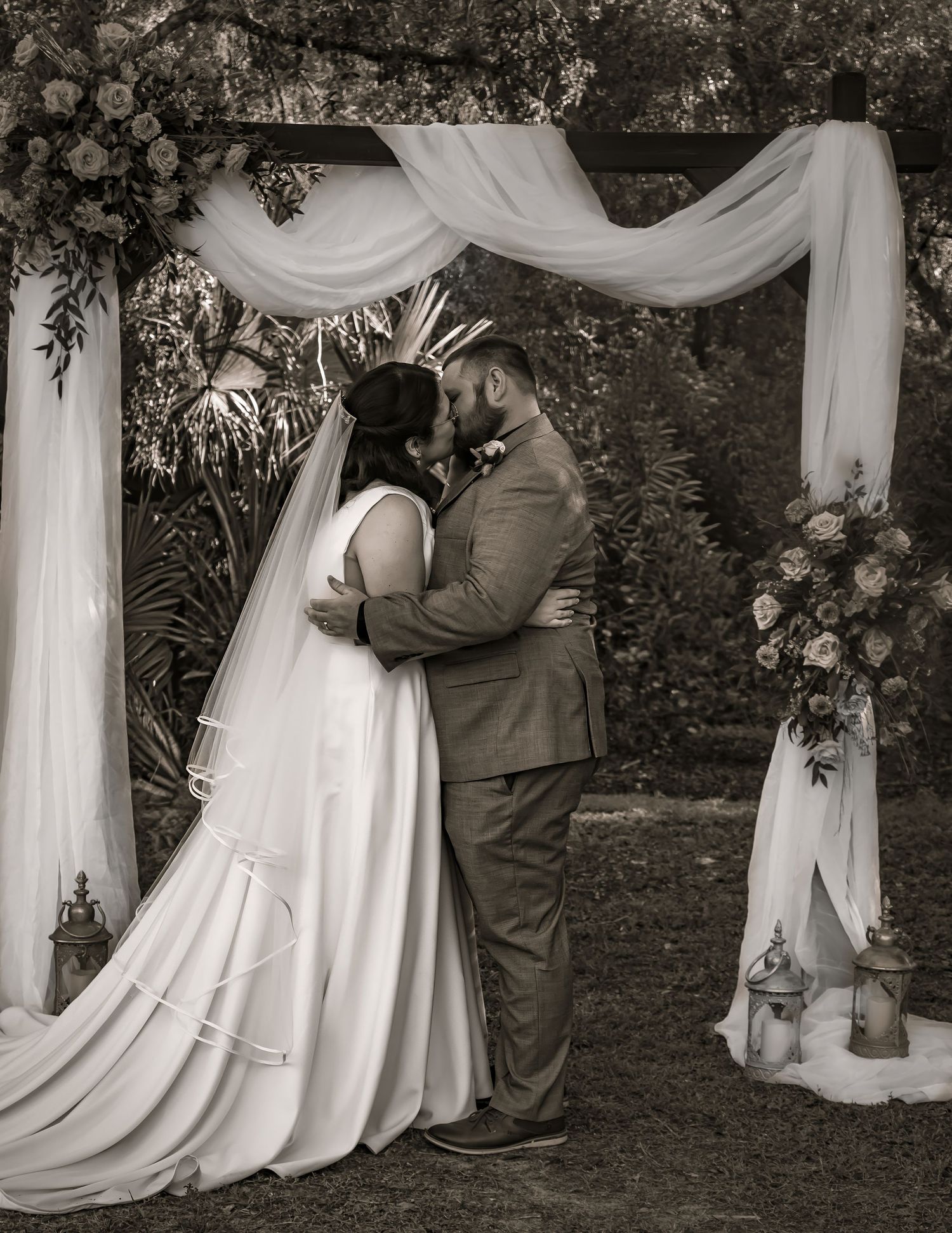 Newlyweds share their first kiss under a draped floral wedding arch in sepia.