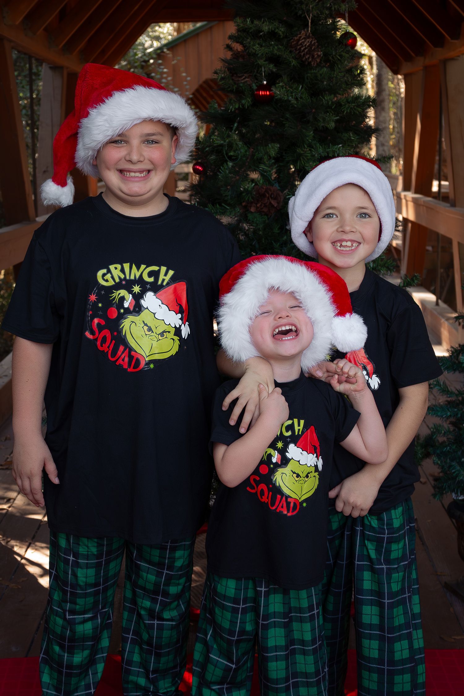 Three children in Grinch Squad shirts and Santa hats smile in front of a Christmas tree.