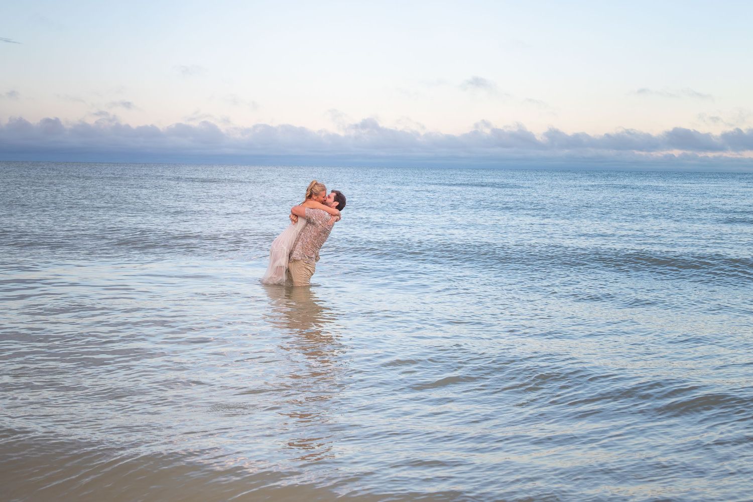 Couple embracing in ocean waves on a sandy beach at sunset, romantic wedding photo.