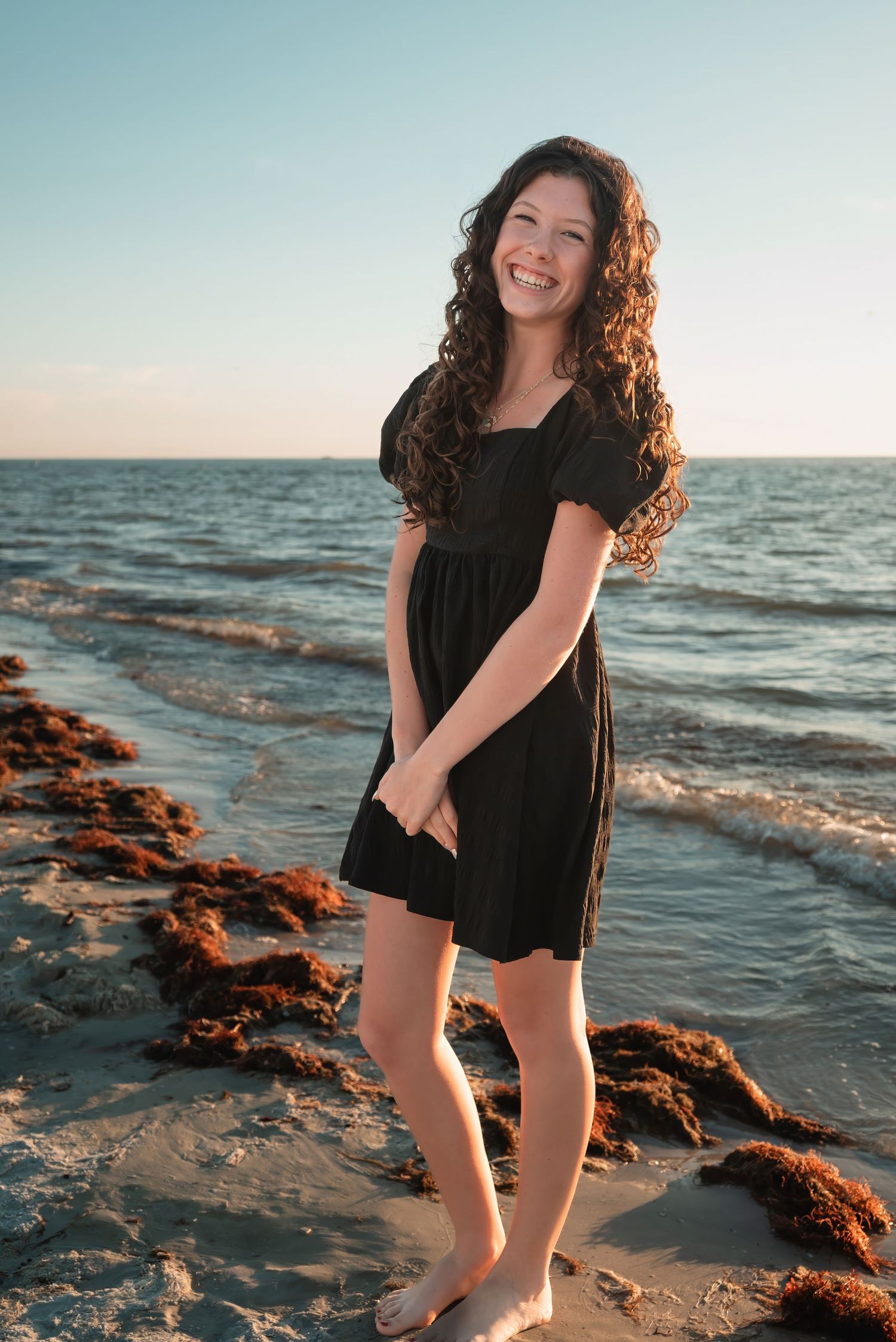 Young woman with curly hair smiling in a black dress standing on a beach at sunset.