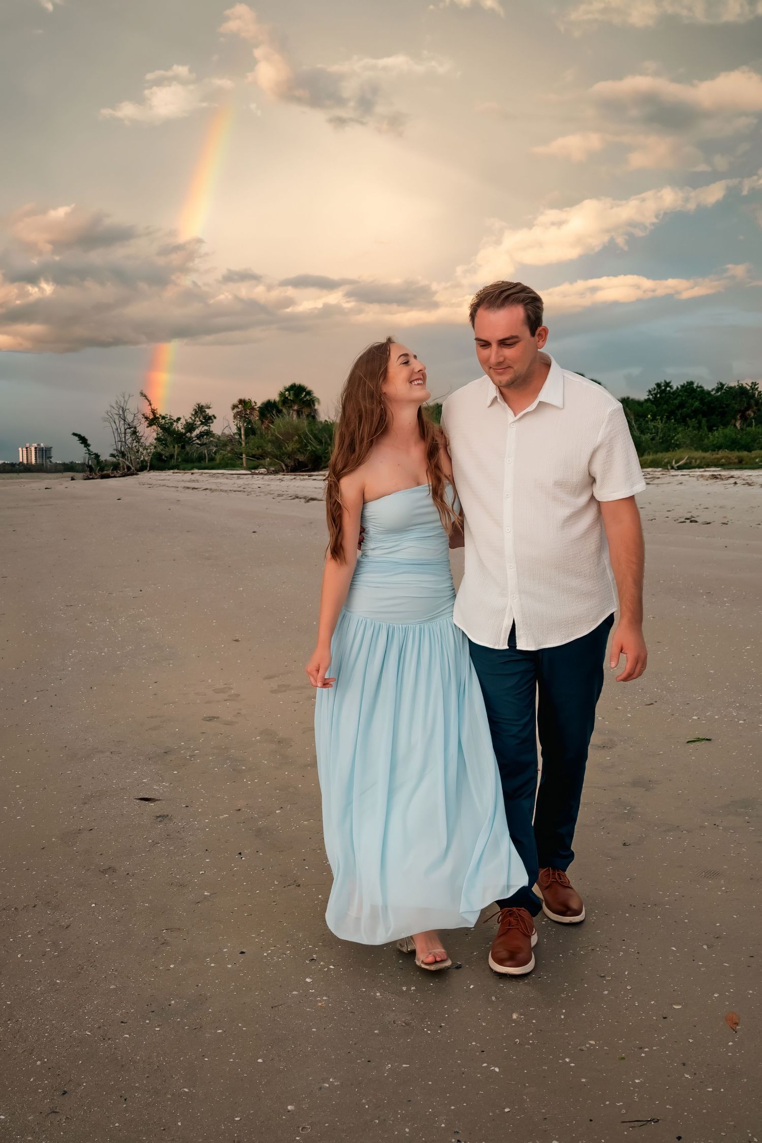 Couple walking on beach in light blue dress and white shirt with rainbow in cloudy sunset sky behind them.