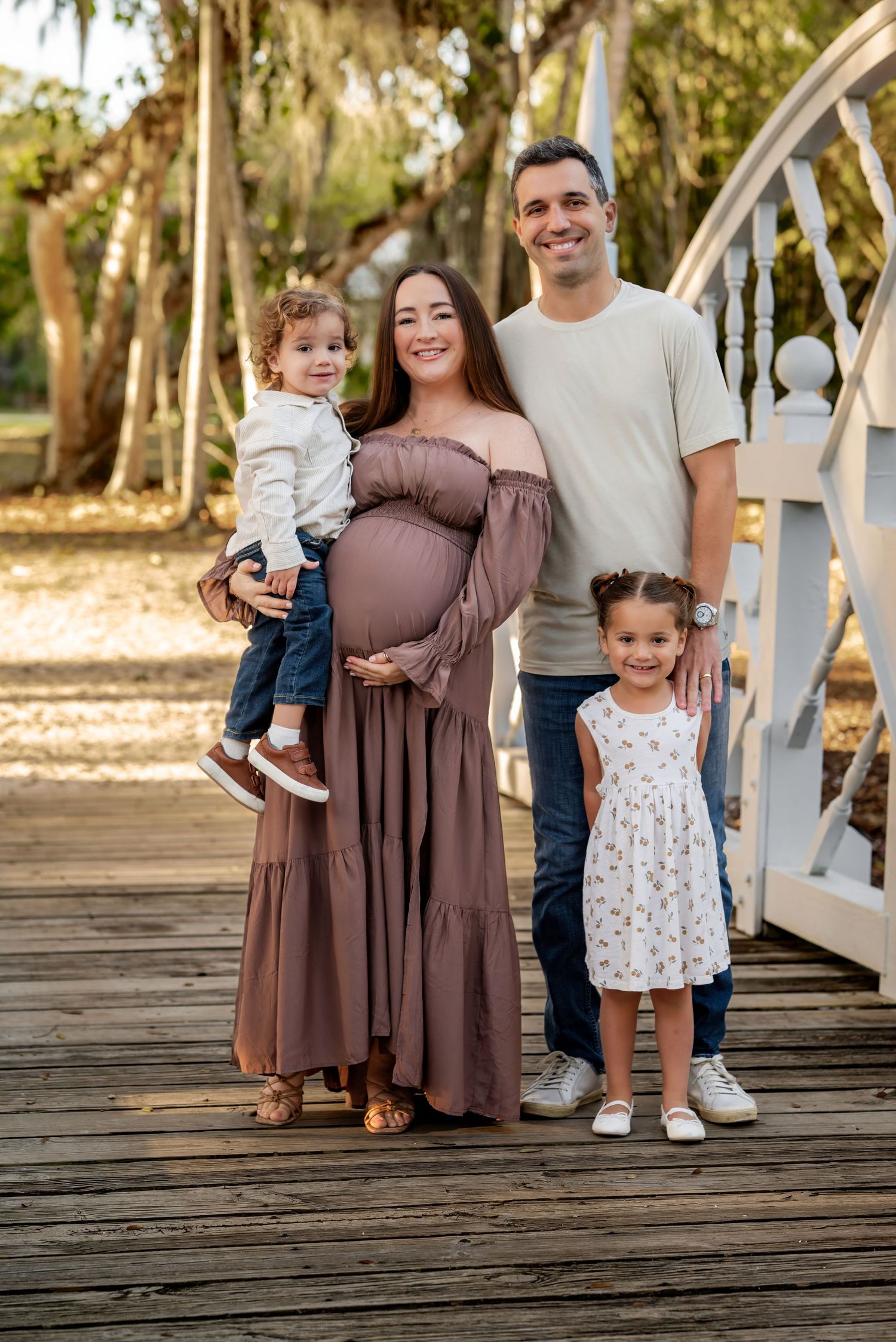 Joyful pregnant family of four poses together on a wooden bridge surrounded by beautiful autumn trees.