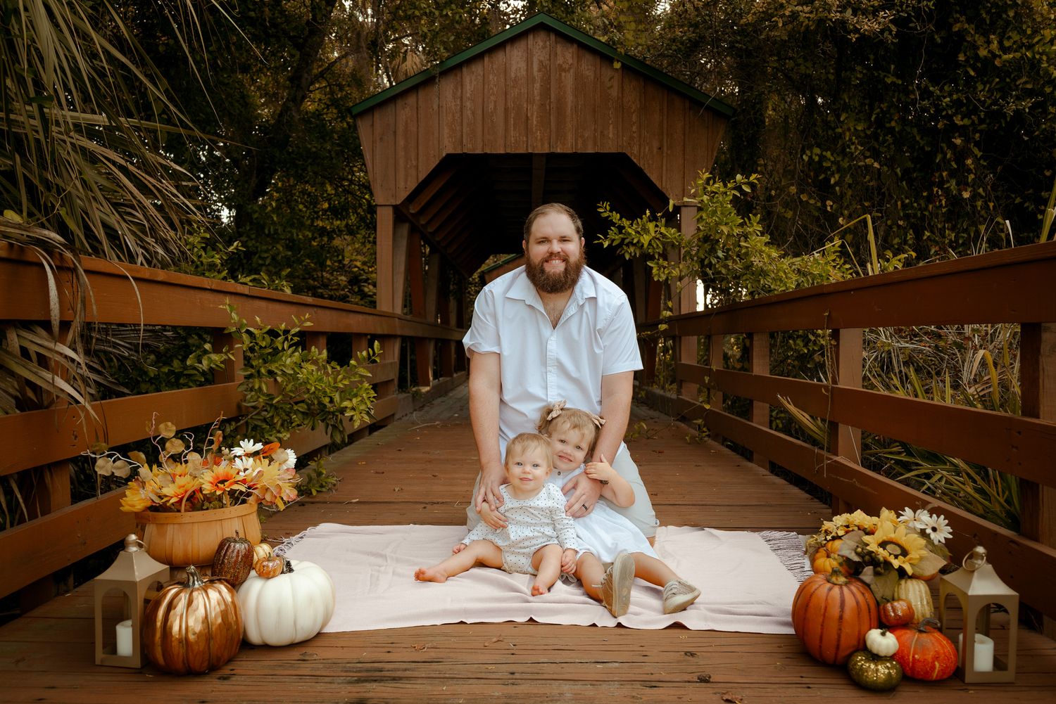 Father poses with two young children on a wooden covered bridge surrounded by fall pumpkins and autumn decorations.