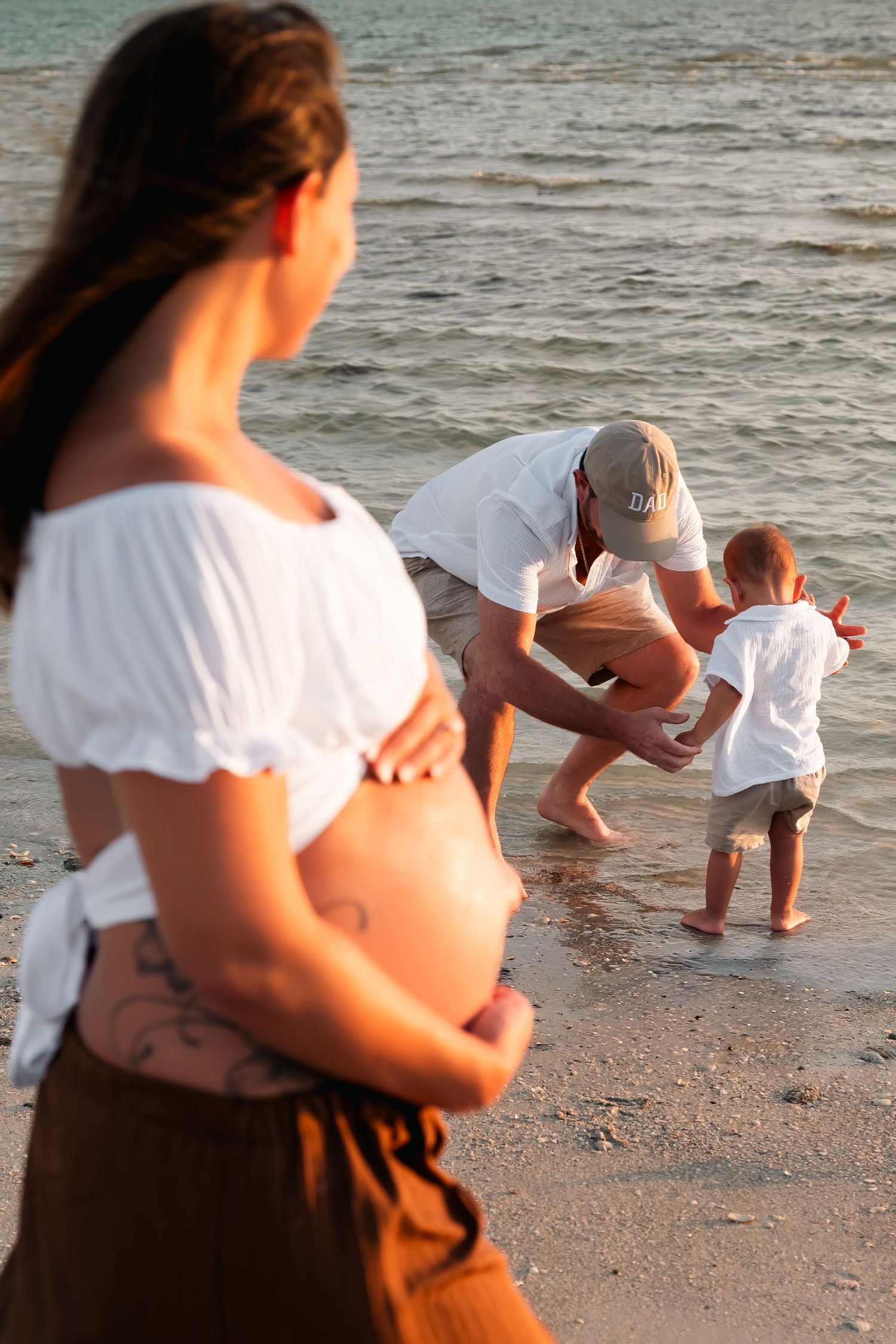 Pregnant woman in white top watches husband and toddler play at ocean's edge during golden hour beach family photo.
