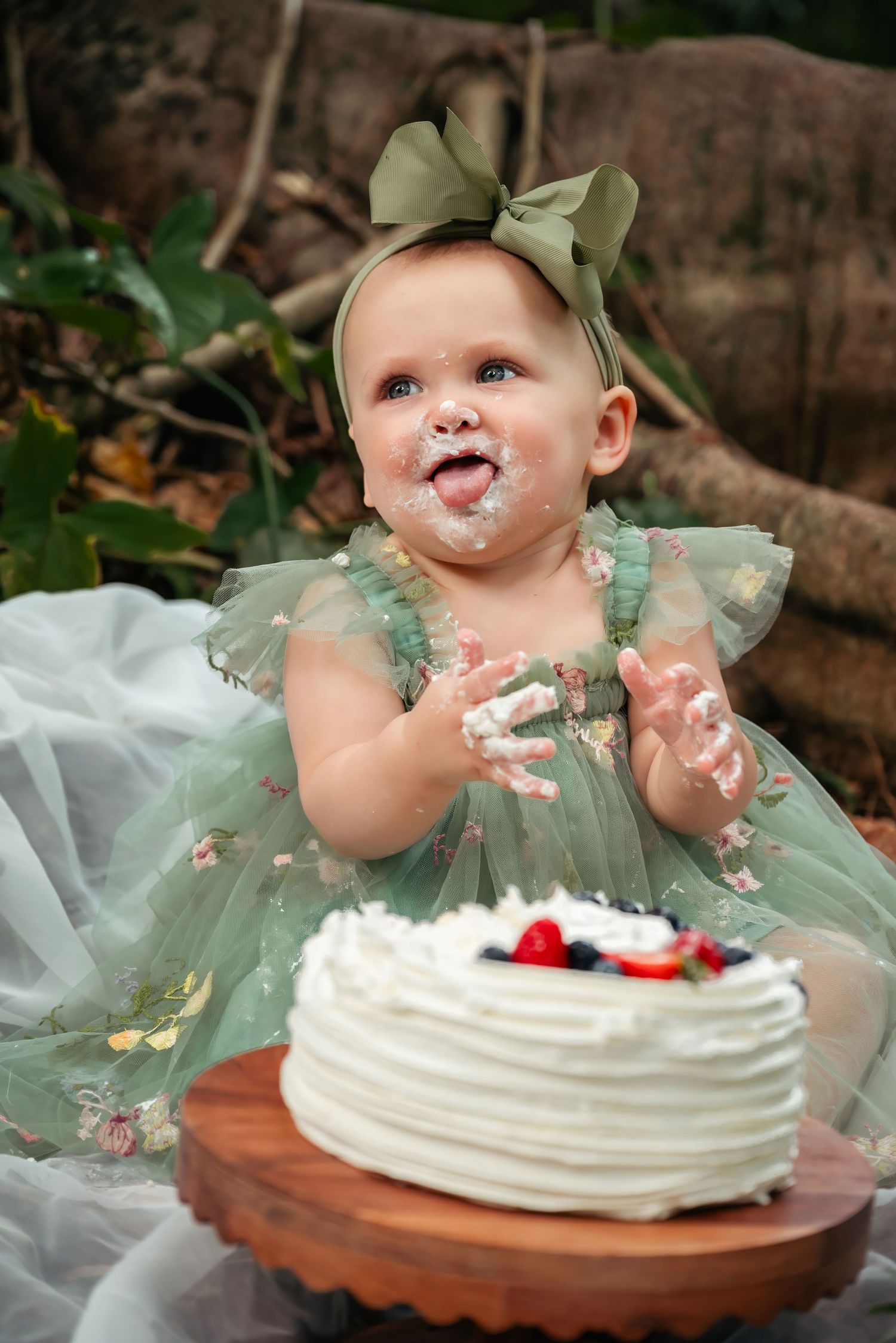 Baby girl in green floral dress clapping with cake on her face during first birthday cake smash photoshoot.