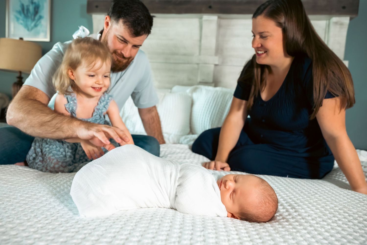 Family of four on bed, parents smiling at swaddled newborn baby while toddler girl reaches out to touch sibling.