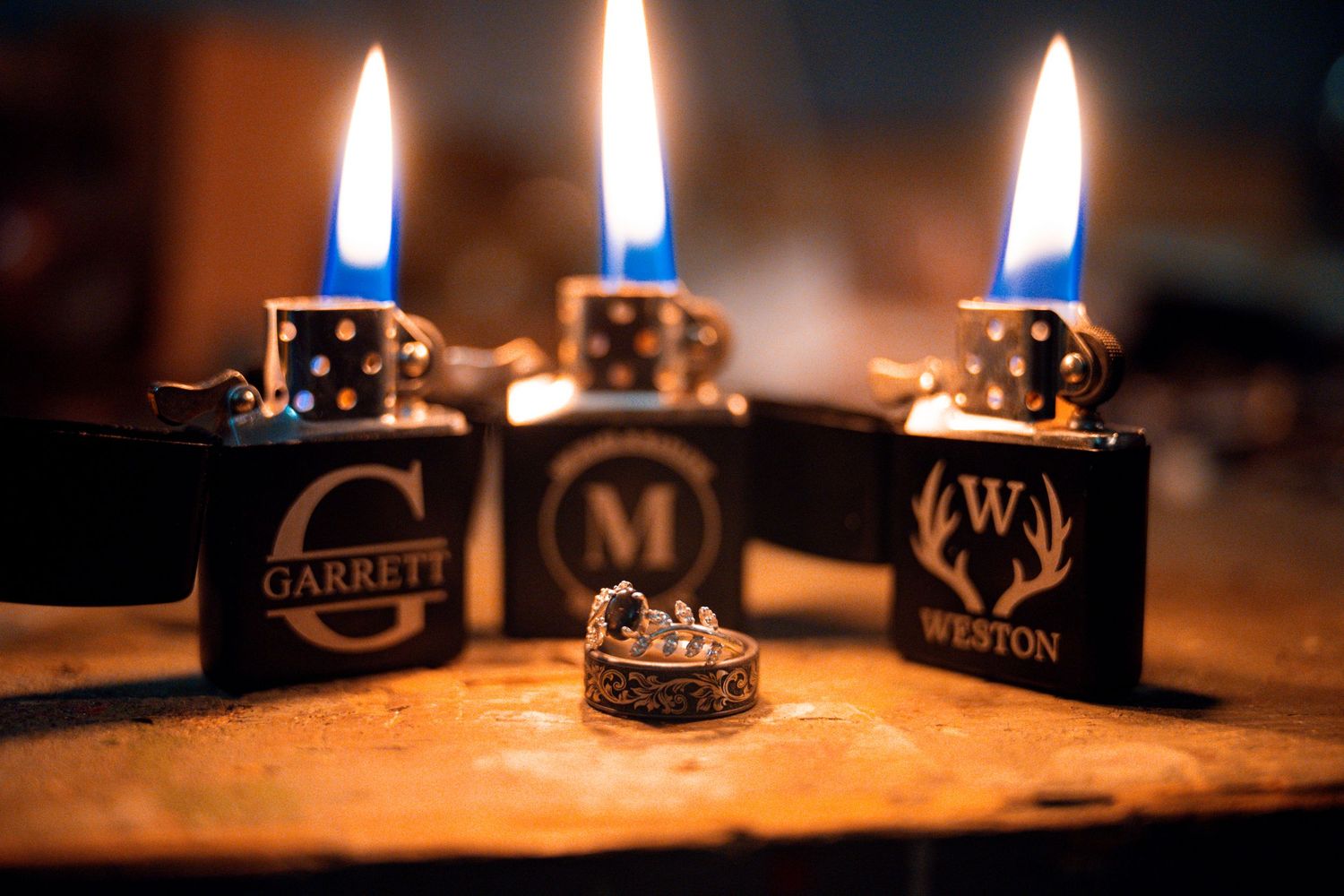 Three personalized engraved Zippo lighters with names Garrett and Weston lit with blue flames beside a diamond ring on a wooden surface.