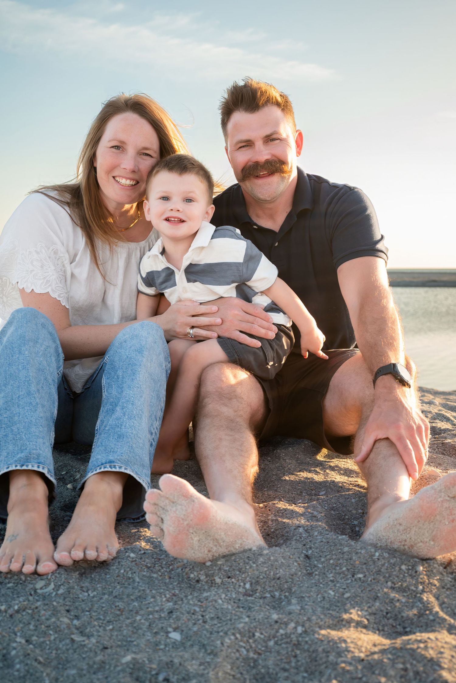 Family of three sitting barefoot on sandy beach, smiling parents with young toddler boy, ocean and blue sky behind them.