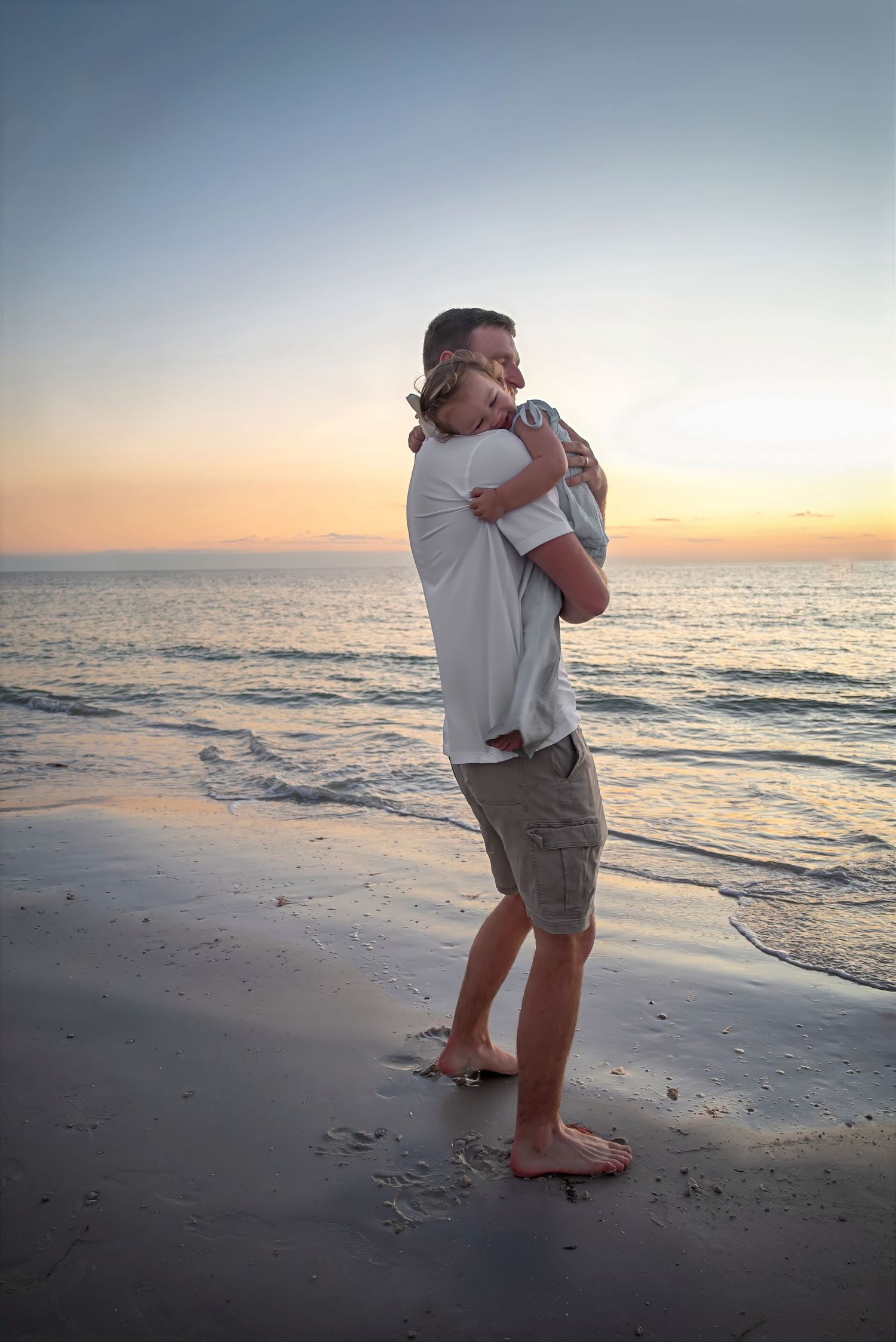 Father holding sleeping child in tender embrace while standing on a beach at golden sunset.