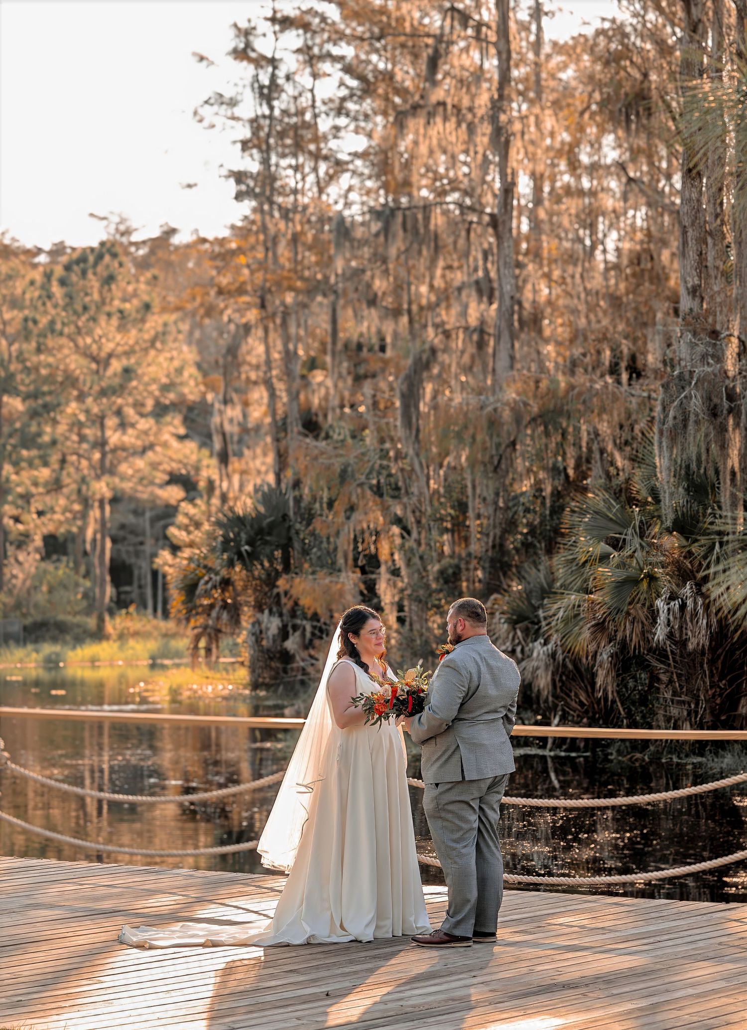 Bride and groom exchange vows on a wooden dock surrounded by cypress trees draped in Spanish moss at golden hour.