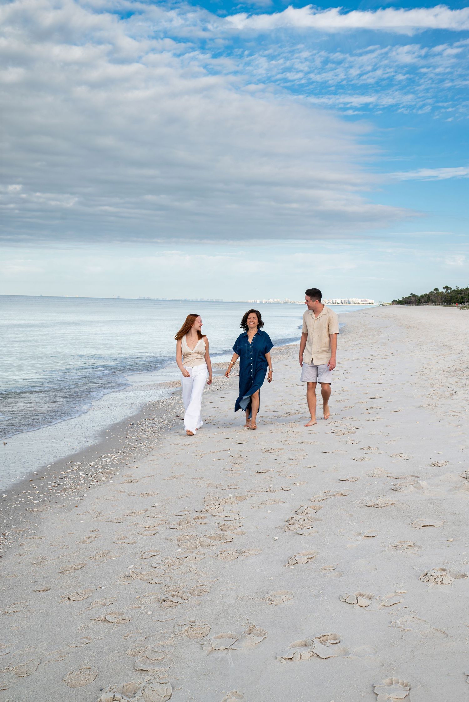 Three family members walking barefoot along a sandy beach under blue sky.