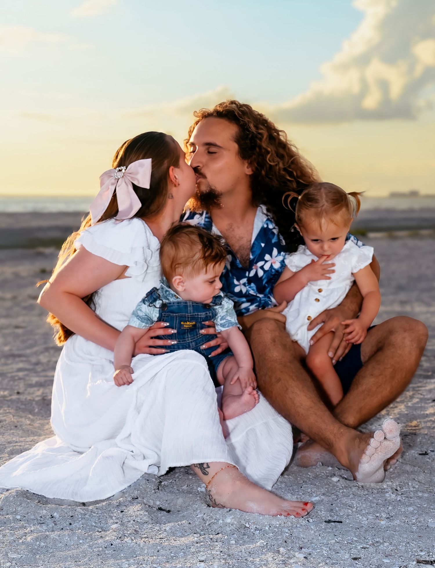 Family of four sitting on beach at sunset, parents kissing while holding two young children.