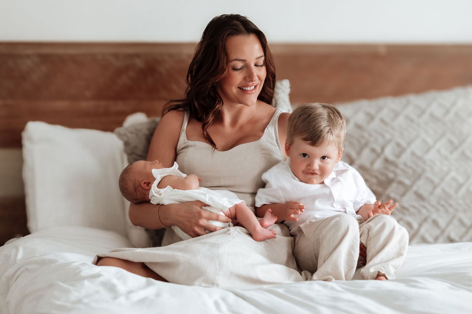 Smiling mother sits on white bed holding newborn while toddler son sits beside her.