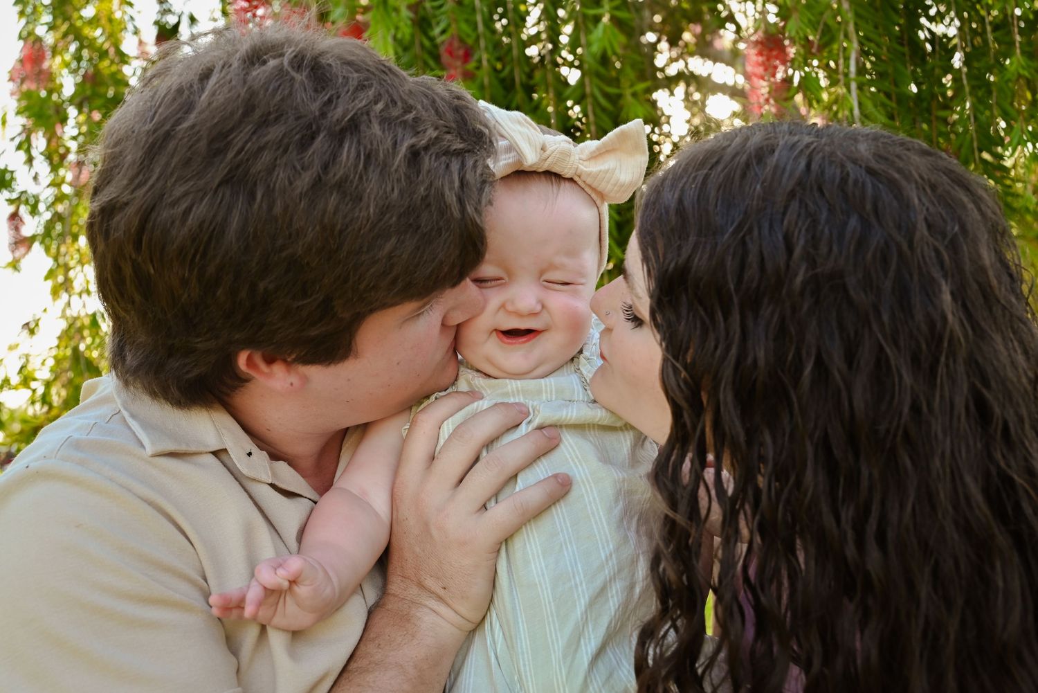 Two parents lovingly kiss their smiling baby on the cheeks outdoors.