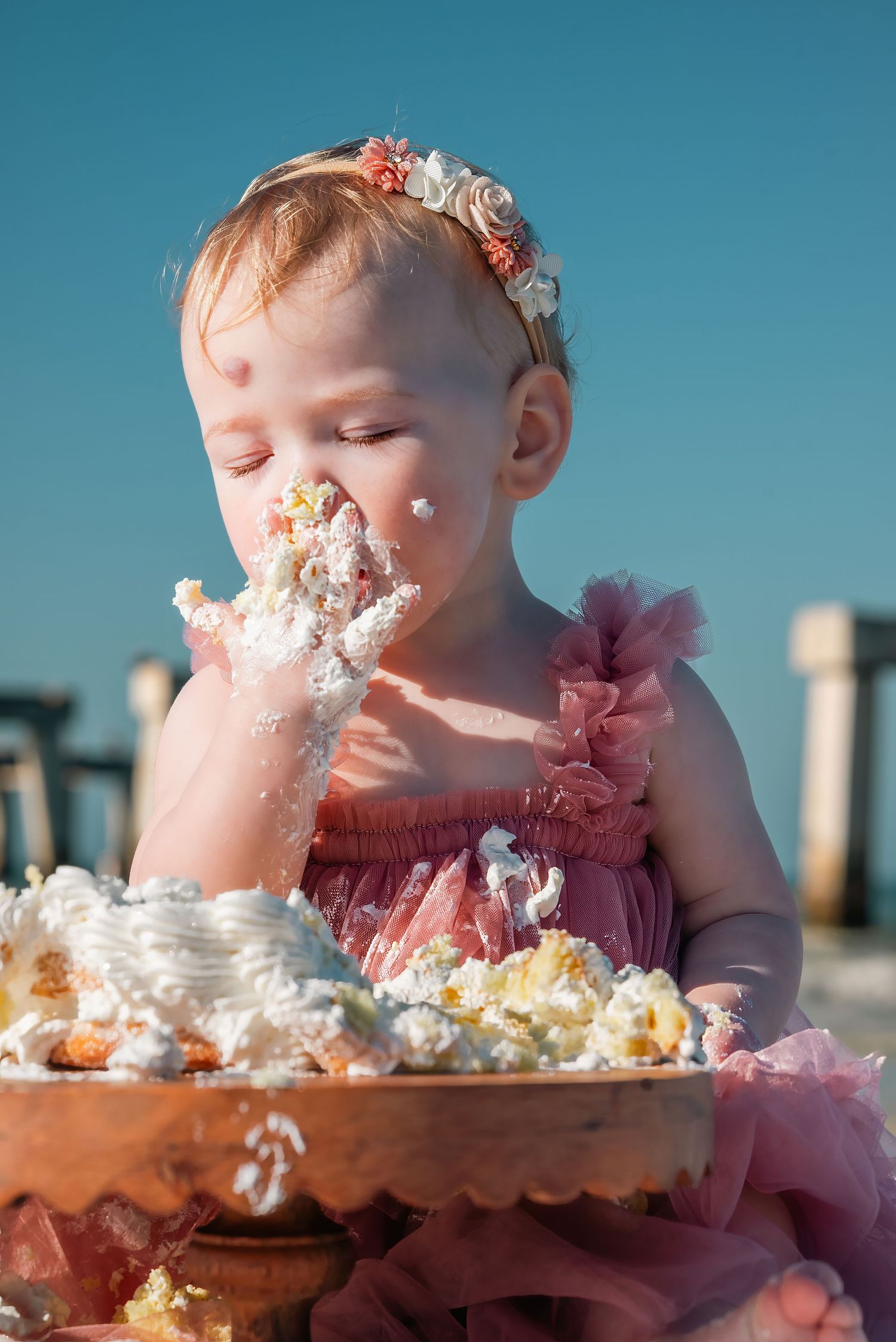 Toddler girl in pink dress enjoying messy cake smash celebration at the beach.