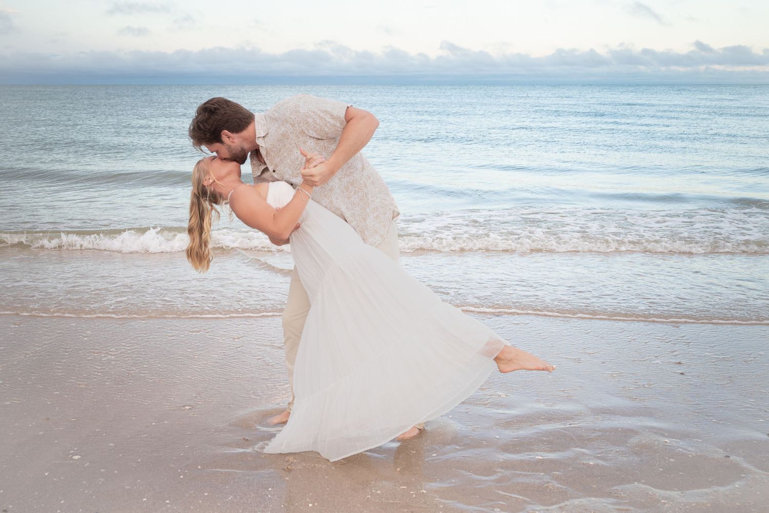 Groom dipping and kissing bride in white wedding dress on sandy beach with ocean backdrop.