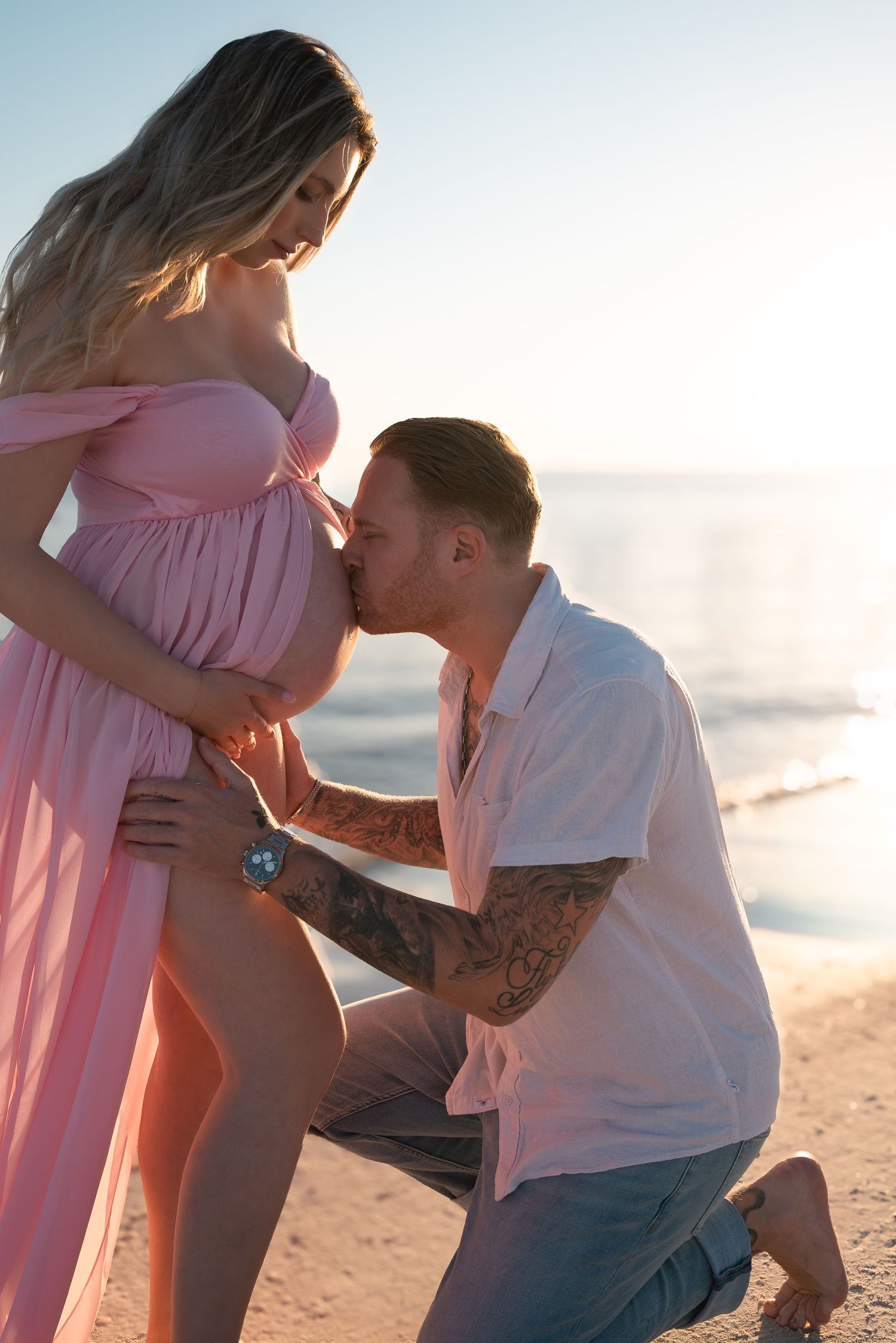 Tattooed man kneeling to kiss pregnant woman's belly on a sunny beach at sunset.