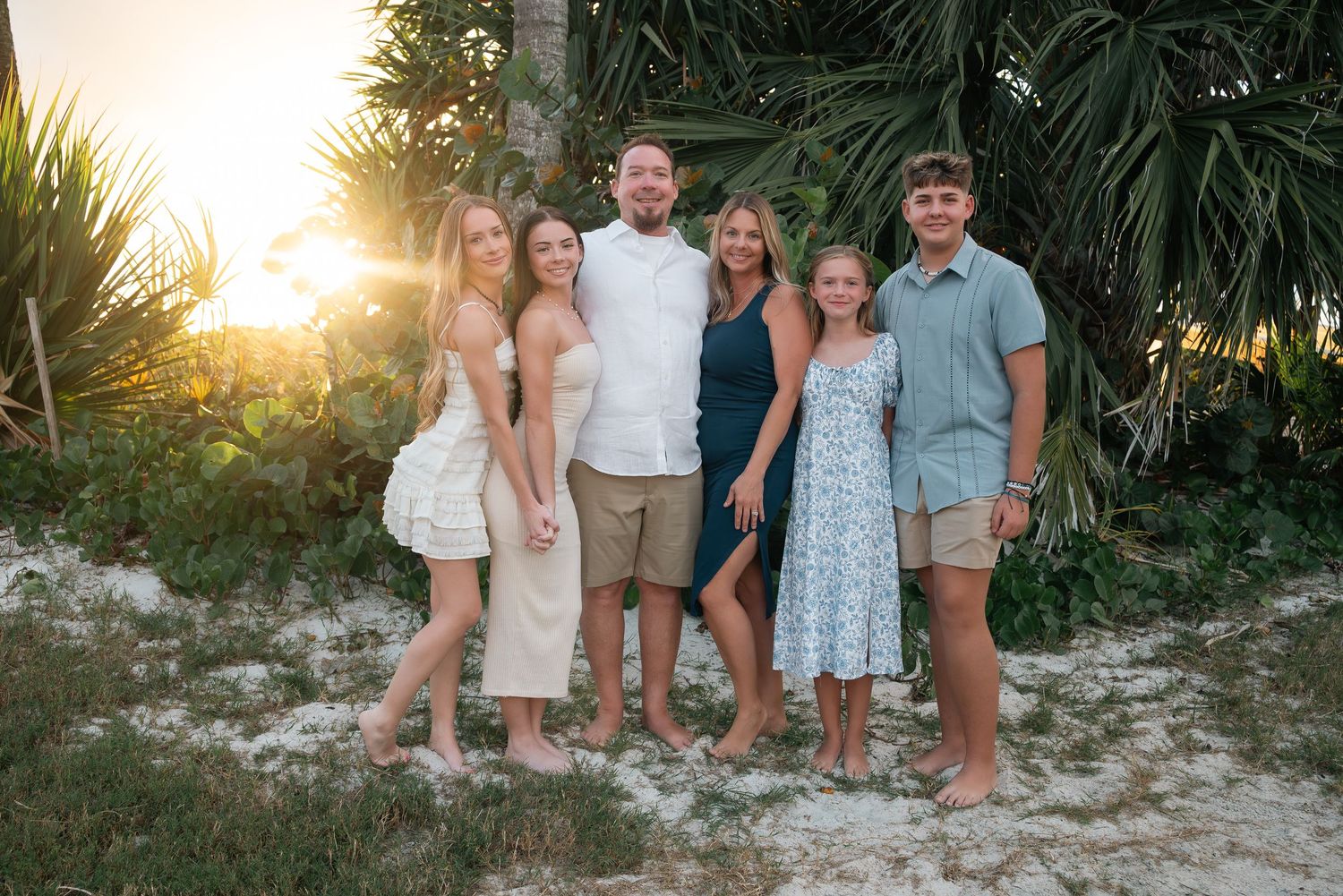Happy family of six poses barefoot among tropical palm trees in warm golden sunset light.