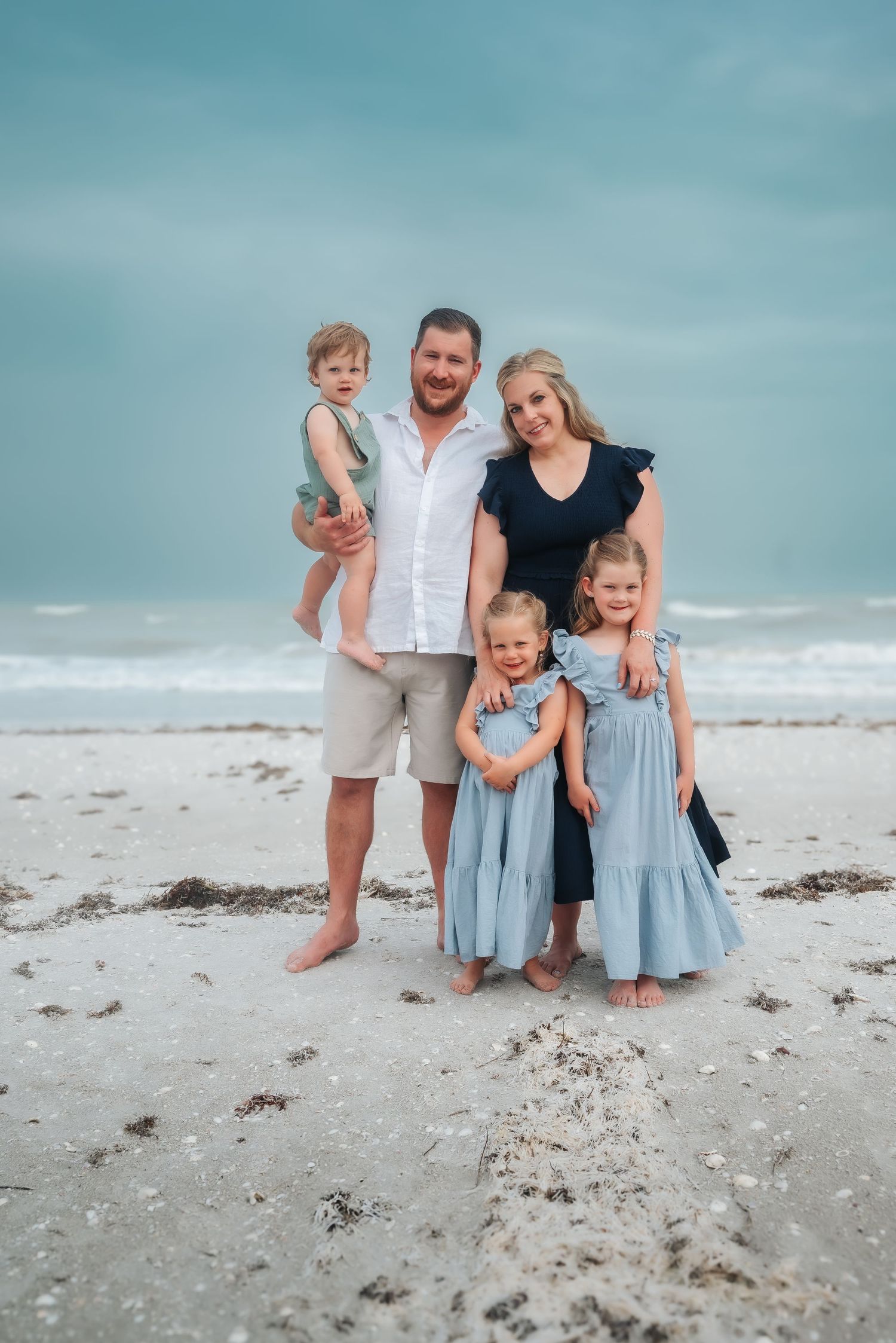 A family of five poses together on a sandy beach under cloudy skies.