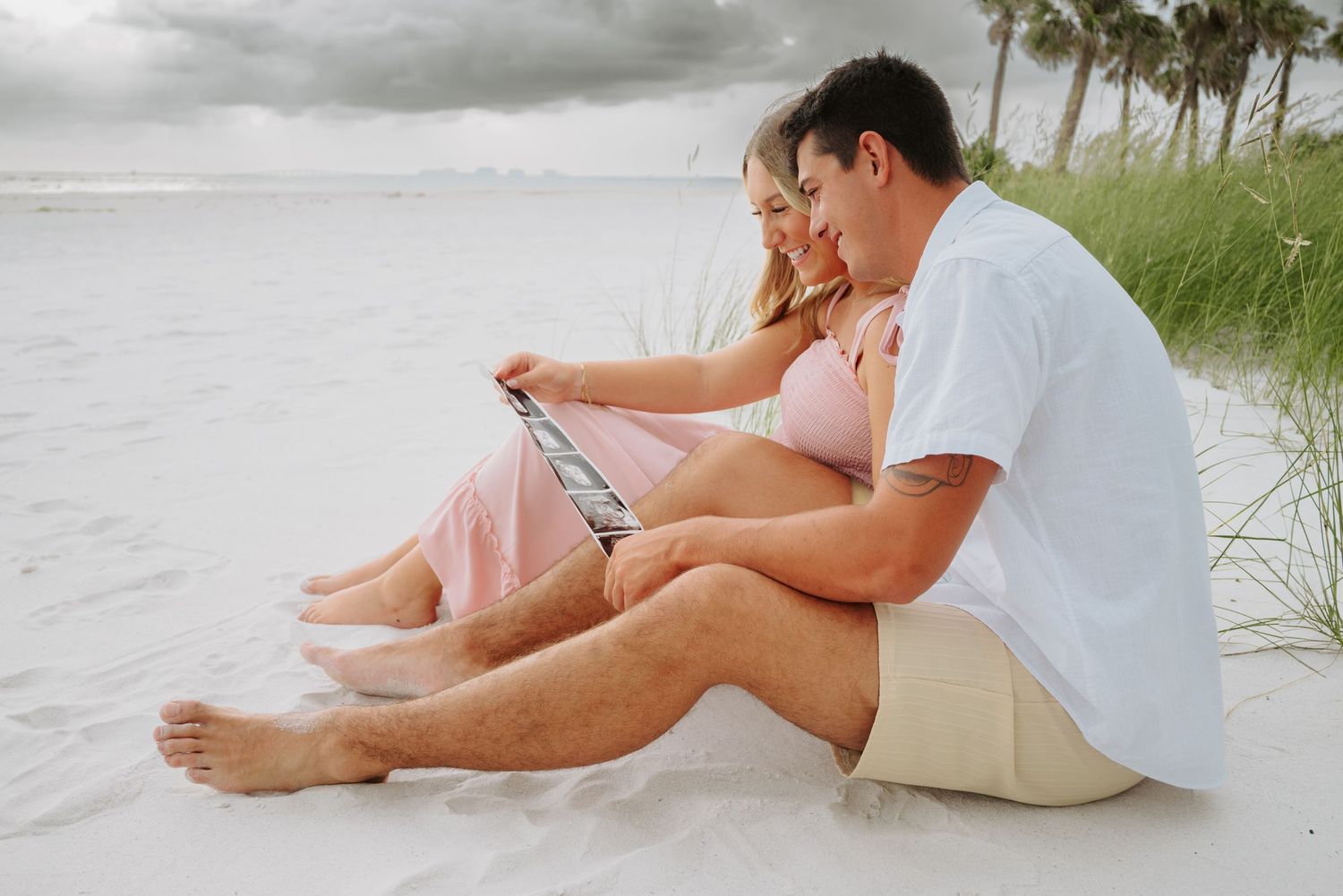 Pregnant couple sitting on white sand beach looking at ultrasound photo together on cloudy day.