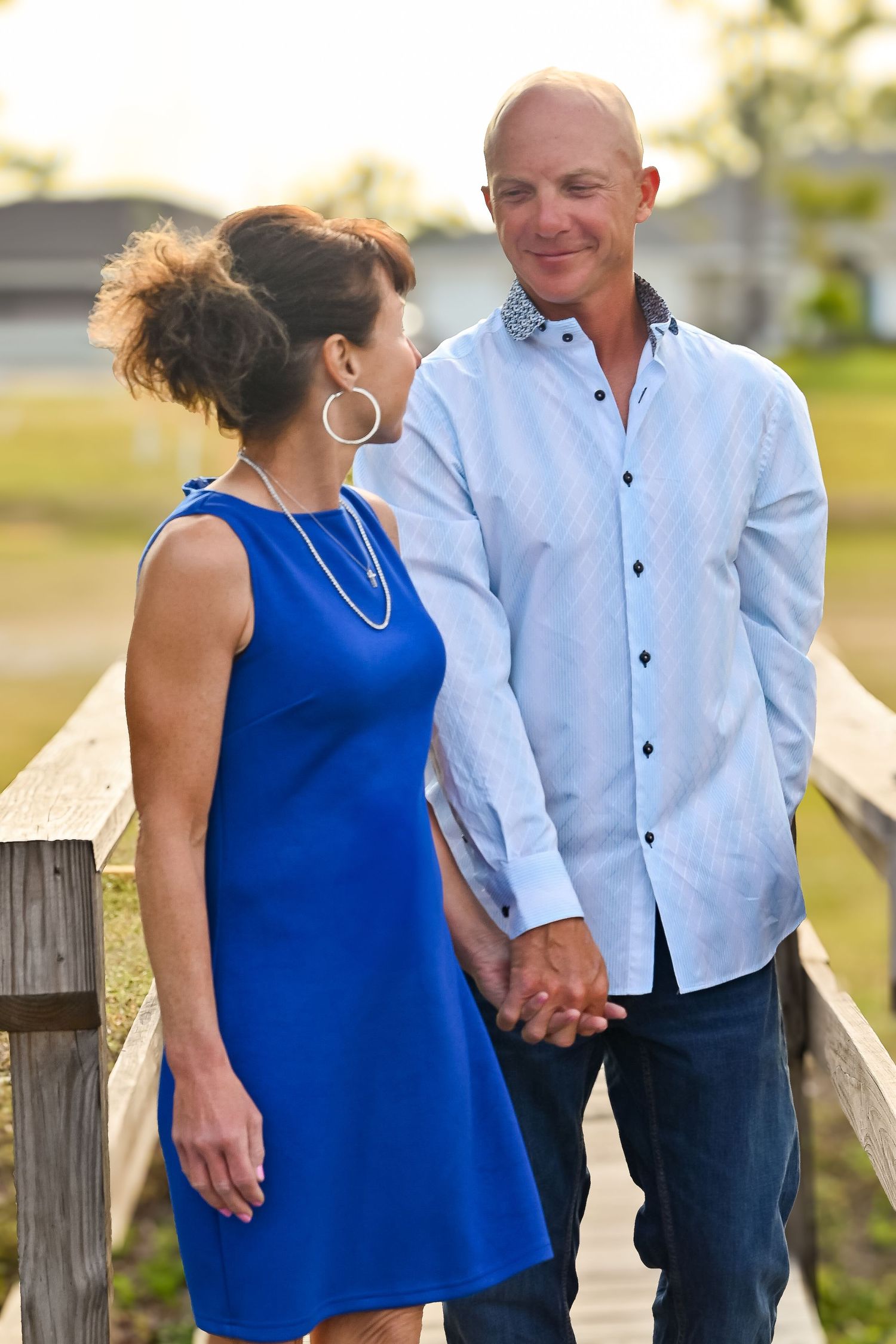 Couple walks hand in hand on wooden bridge, man gazes at woman in blue dress.