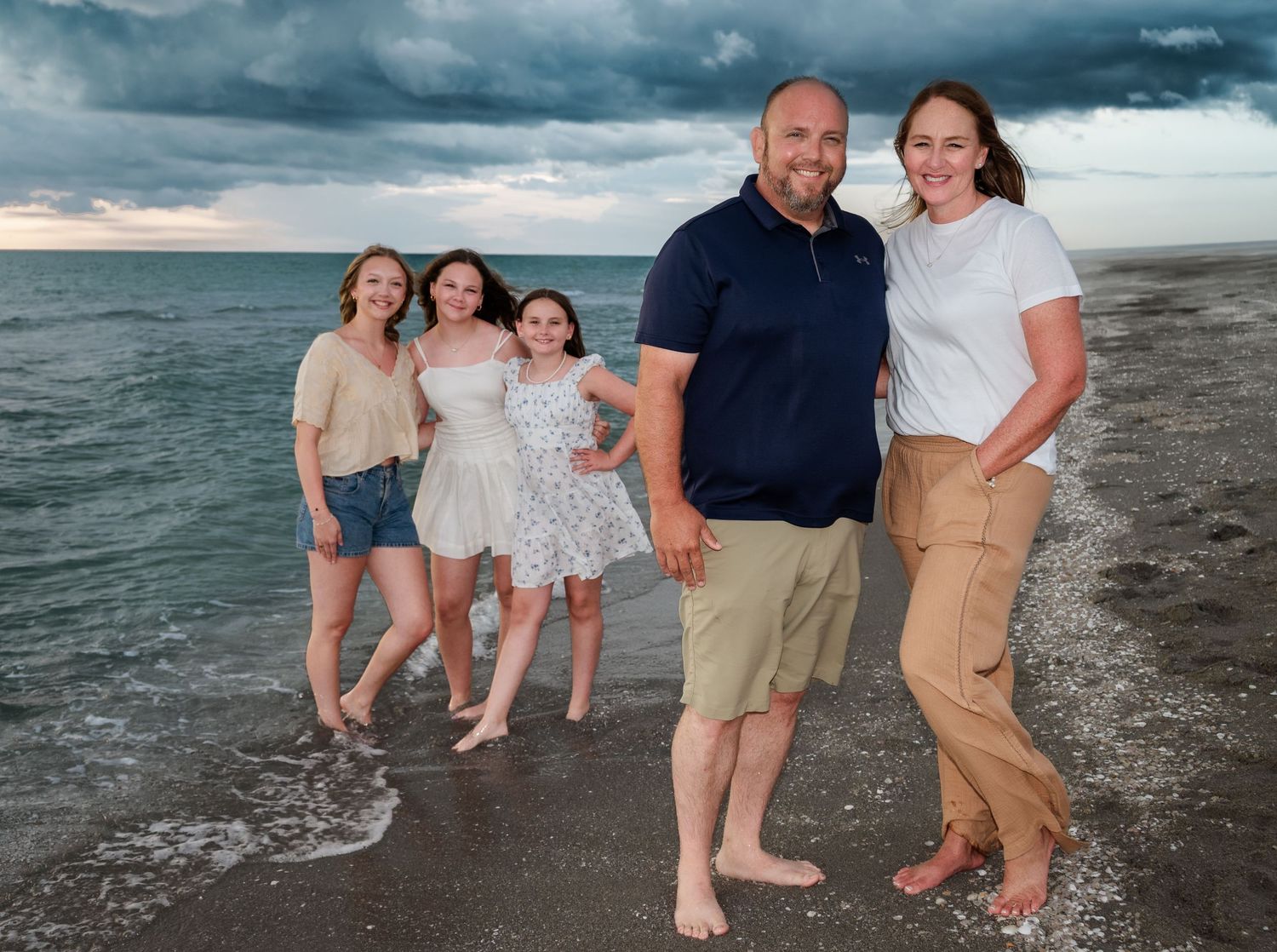 Family of six posing together on beach shoreline with stormy sky backdrop.