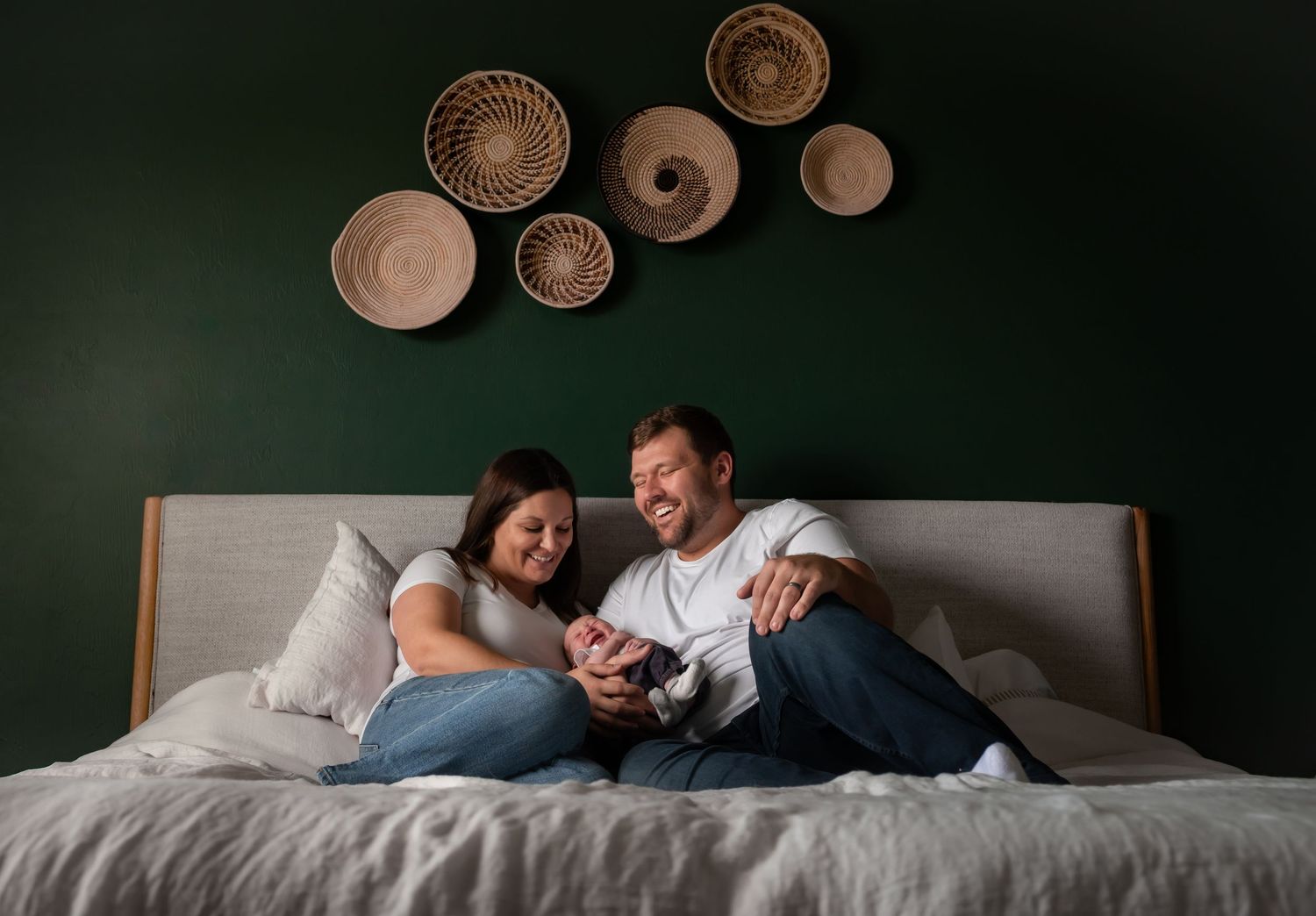 Happy couple sitting on bed with newborn baby, green wall with woven basket decor behind them.