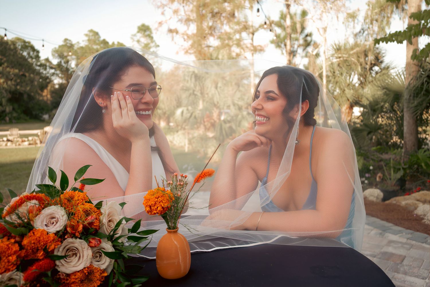 Two brides share a joyful moment under a veil outdoors with orange and white wedding flowers nearby.