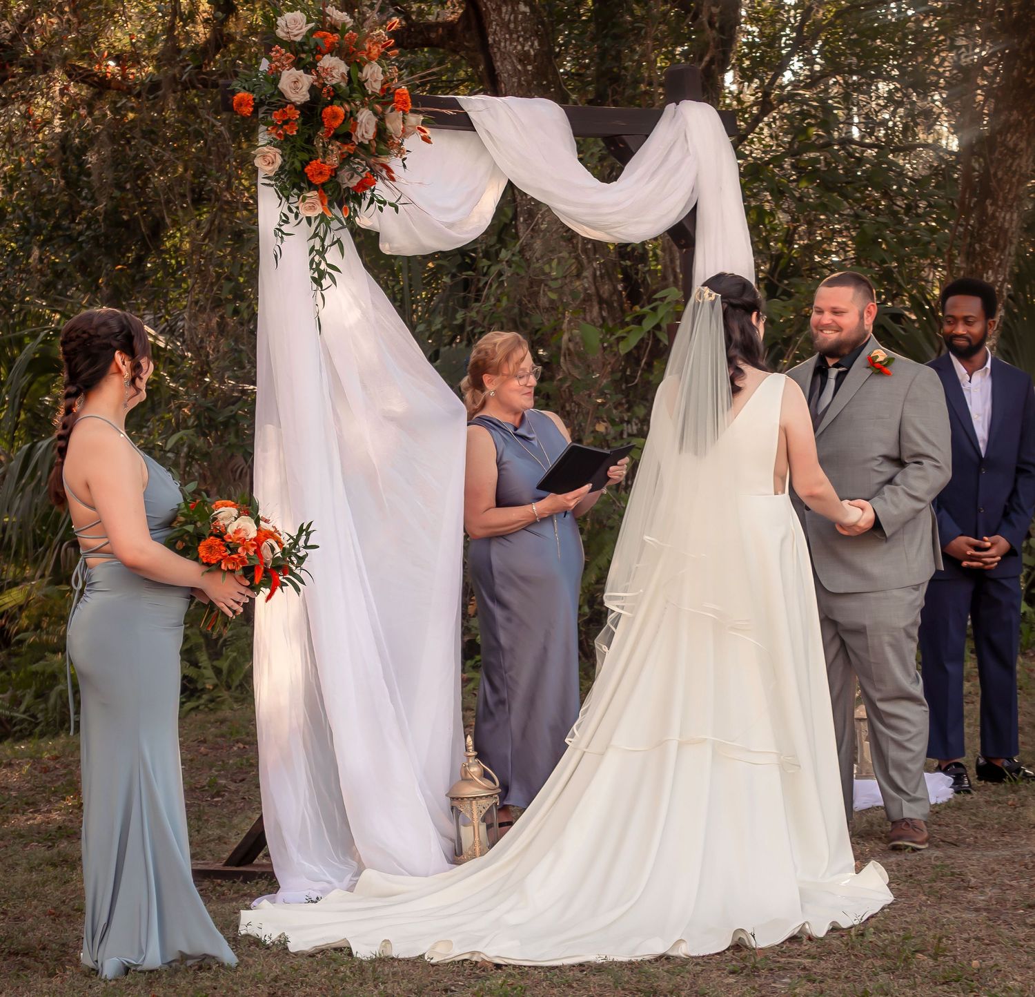 Bride in flowing white gown stands at floral wedding arch with groom and officiant during ceremony.